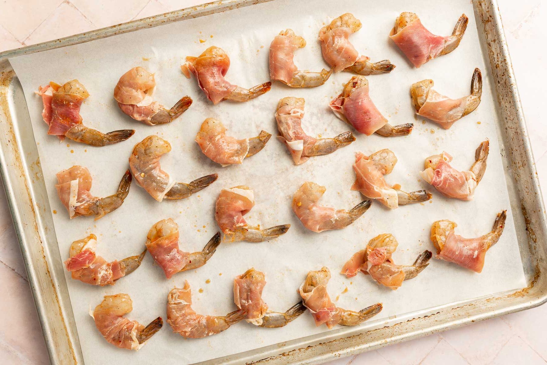 Raw shrimp wrapped in prosciutto and placed in rows on a parchment paper lined baking sheet before baking in the oven.