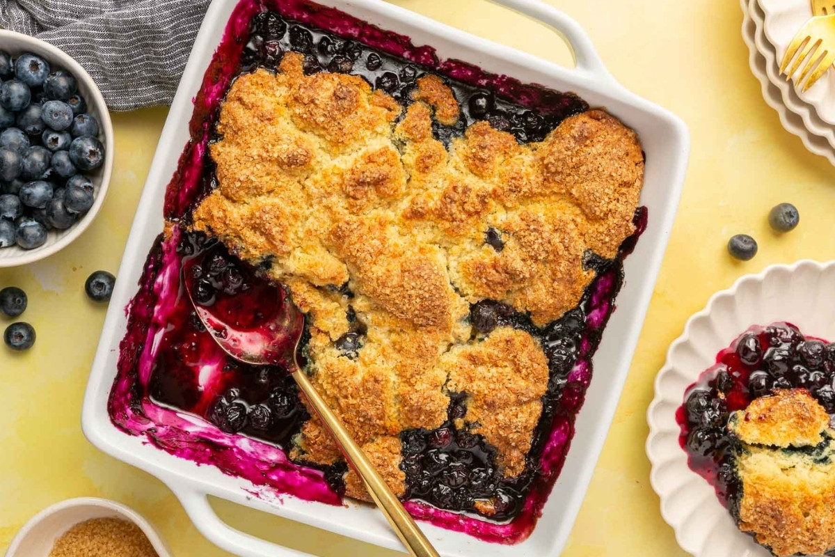 A square white baking dish filled with gluten free blueberry cobbler, topped with golden-brown biscuit crust. A spoon rests in the dish, and a serving is plated nearby. Fresh blueberries and a bowl of sugar are also visible.
