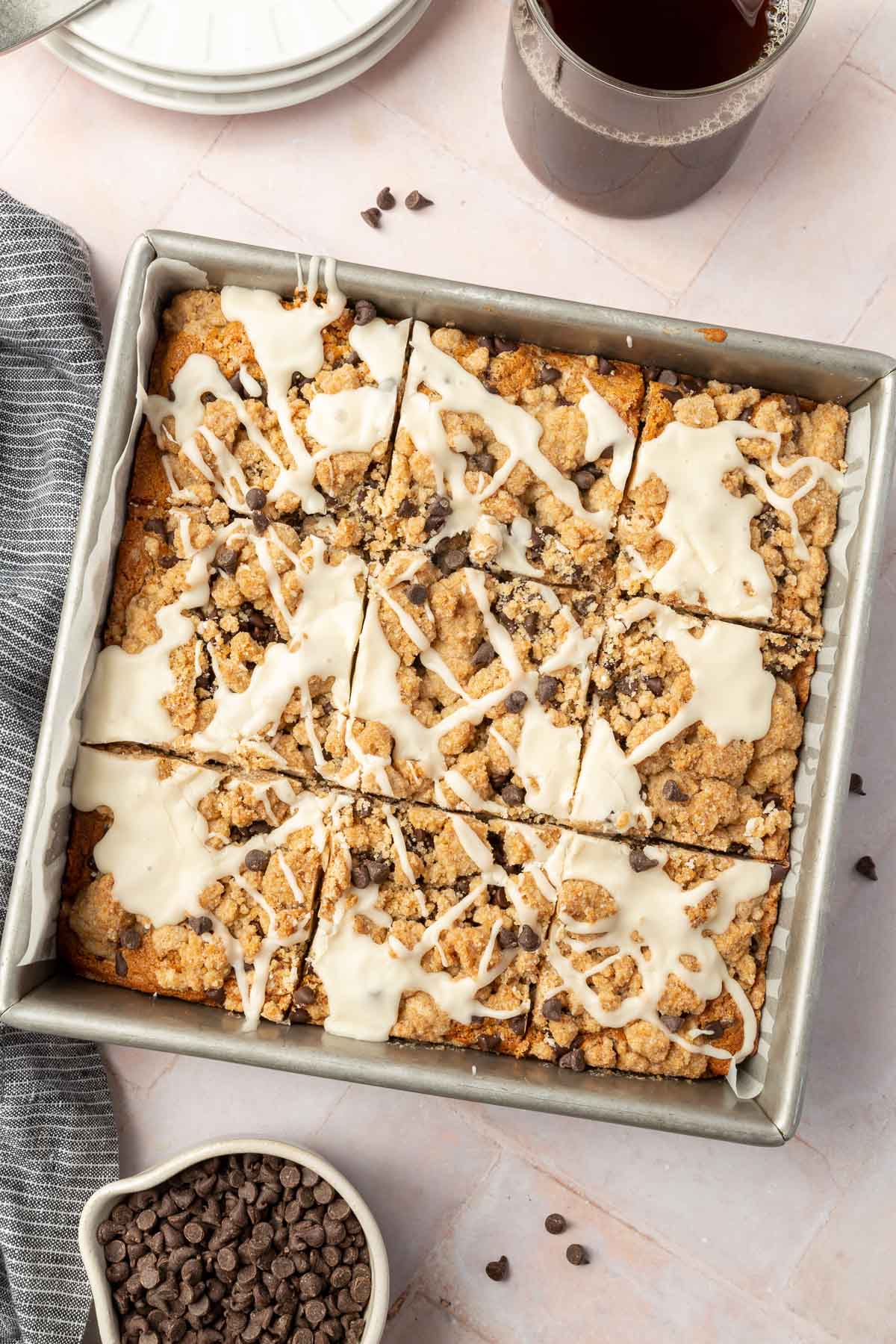 An overhead view of gluten free chocolate chip coffee cake in a square metal baking pan cut into 9 equal slices with a bowl of mini cocolate chips and a cup of coffee off to the side.