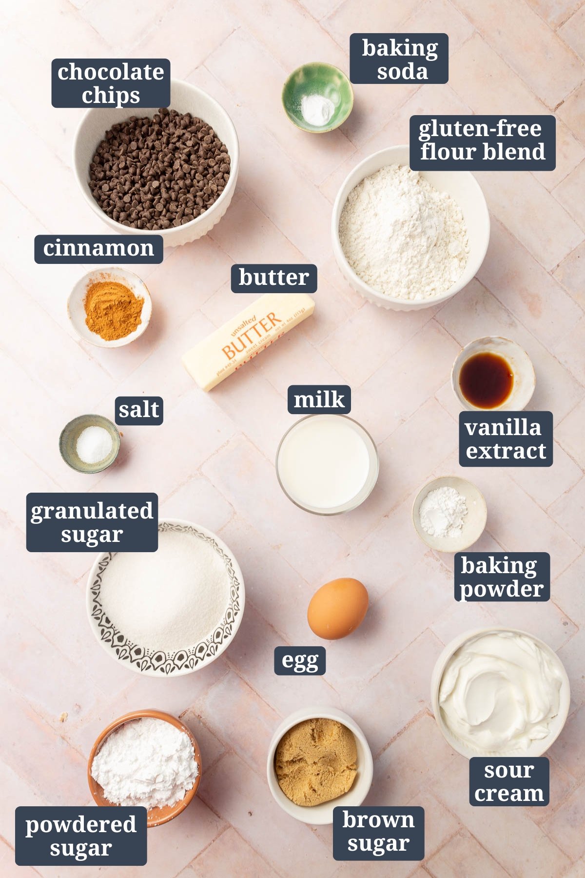 An overhead view of ingredients in small bowls to make gluten free chocolate chip coffee cake with text overlays over each ingredient, including chocolate chips, baking soda, gluten-free flour blend, cinnamon, butter, milk, vanilla, salt, granulated sugar, egg, baking powder, powdered sugar, brown sugar and sour cream.