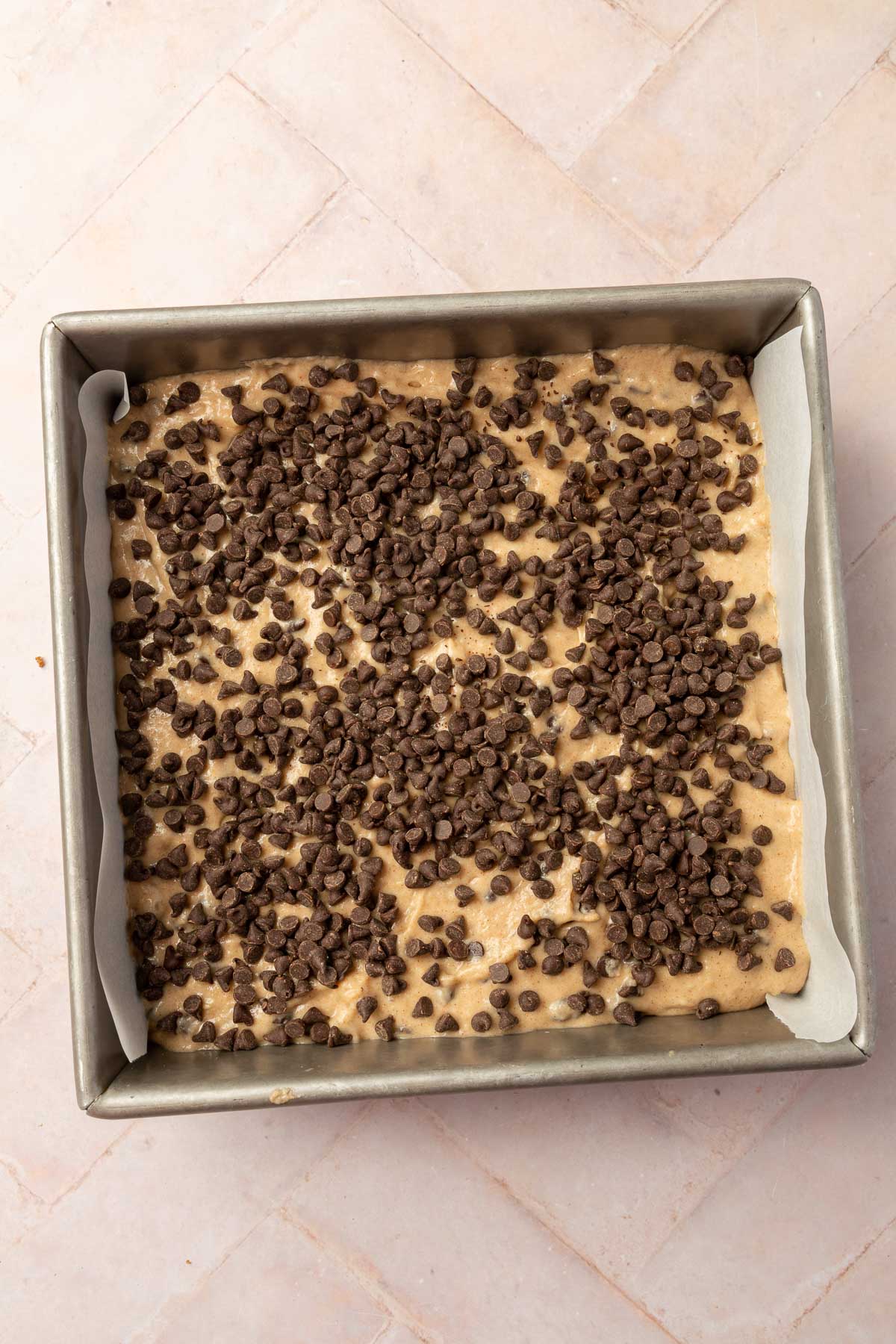 A square metal baking dish filled with gluten-free chocolate chip coffee cake batter topped with additional mini chocolate chips.