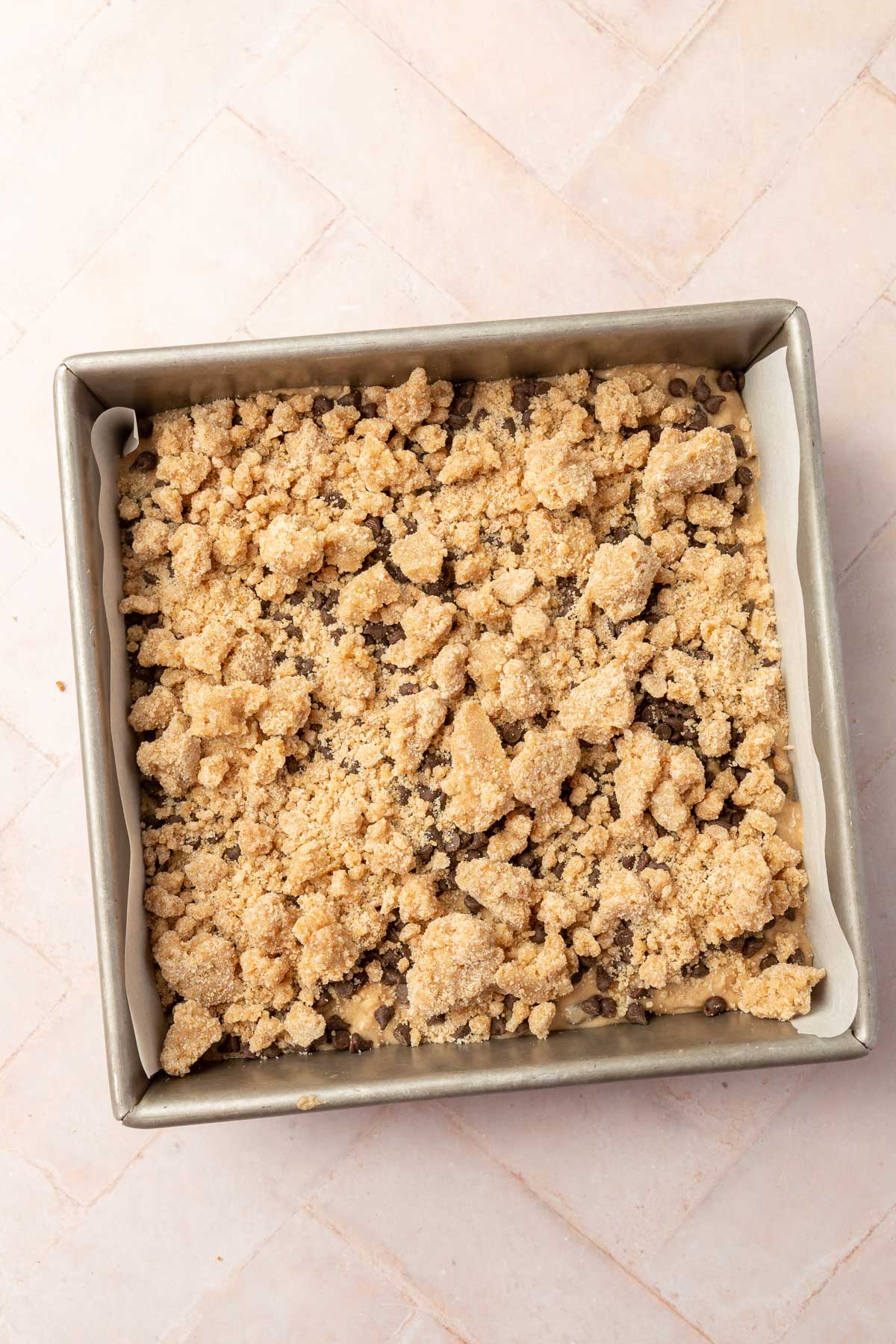 A square metal baking dish with gluten-free chocolate chip coffee cake batter topped with a gluten-free streusel and mini chocolate chips before baking in the oven.