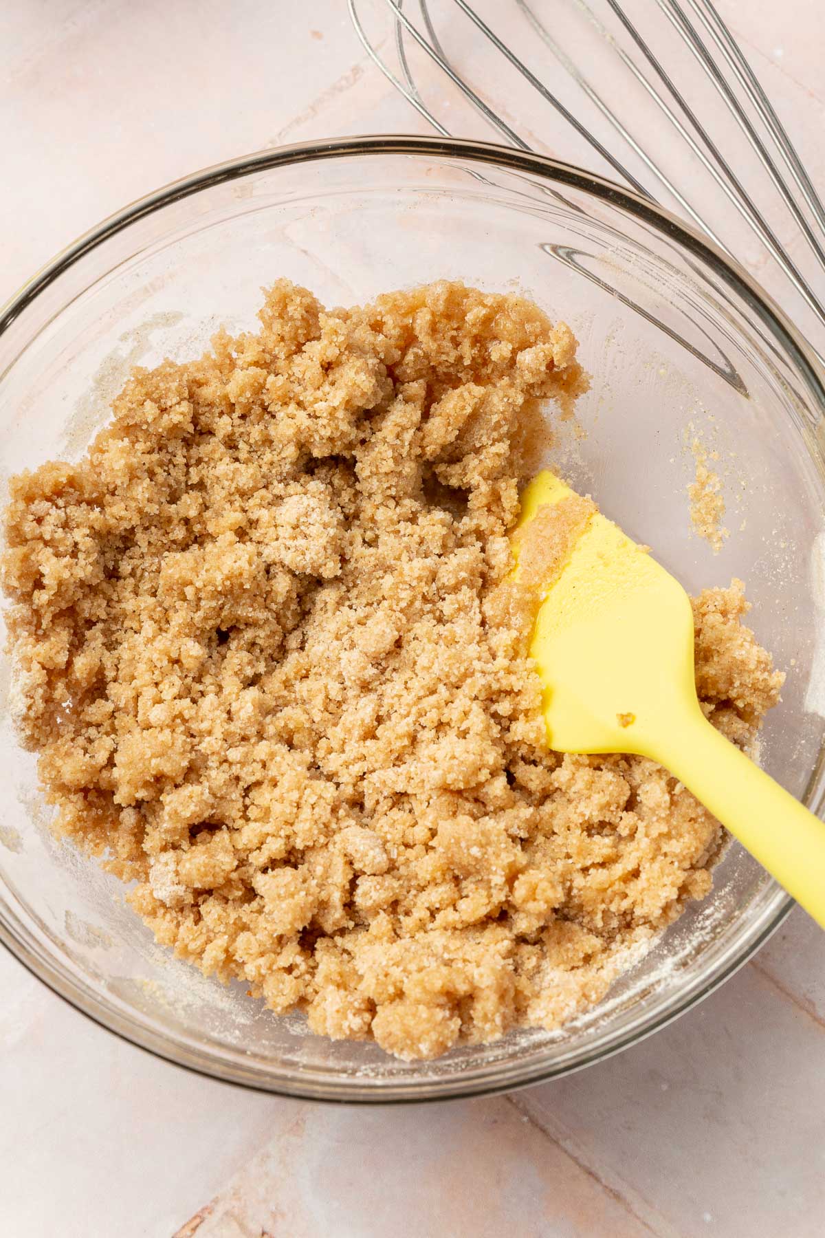 A glass mixing bowl with gluten-free streusel in it with a yellow rubber spatula.