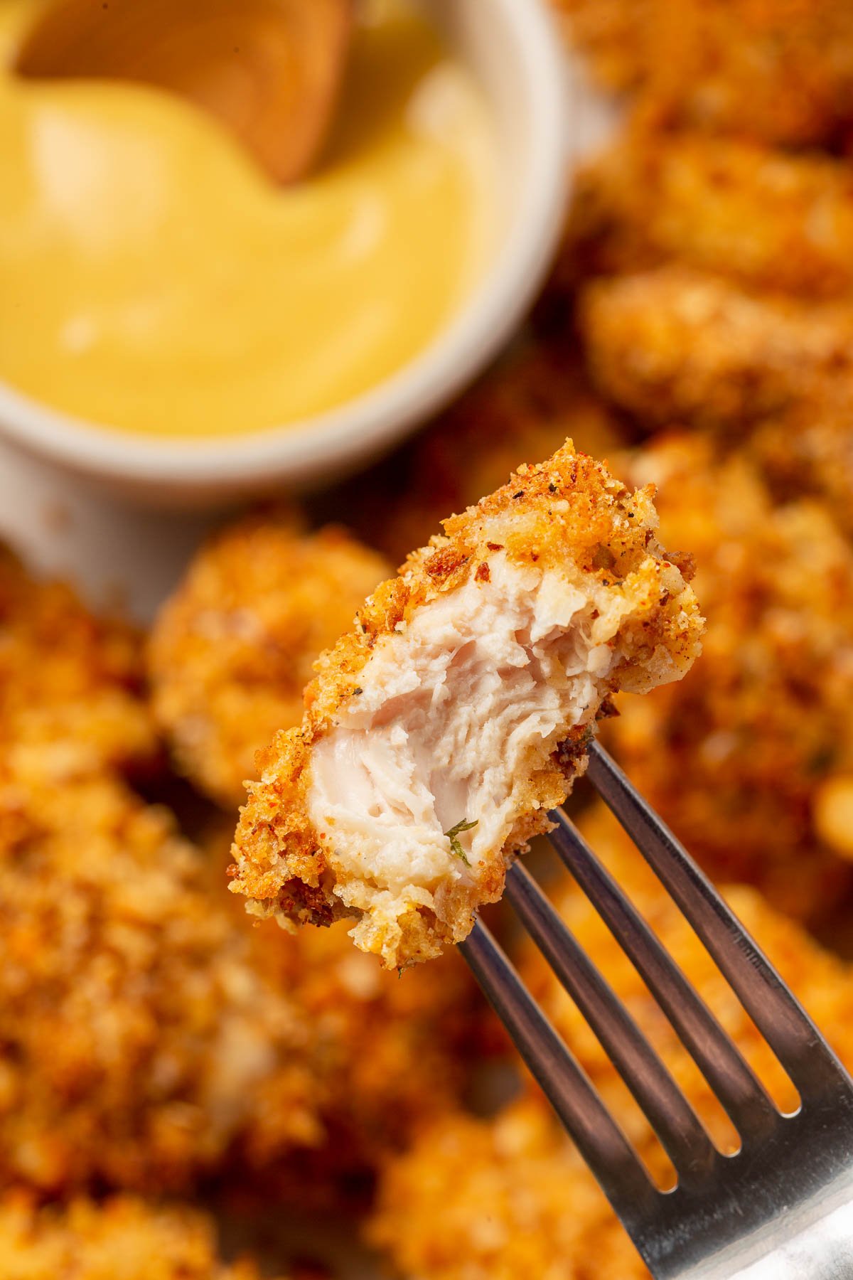 A chicken nugget on a fork that has a bite taken out of it over a platter of additional chicken nuggets with a side of honey mustard sauce.
