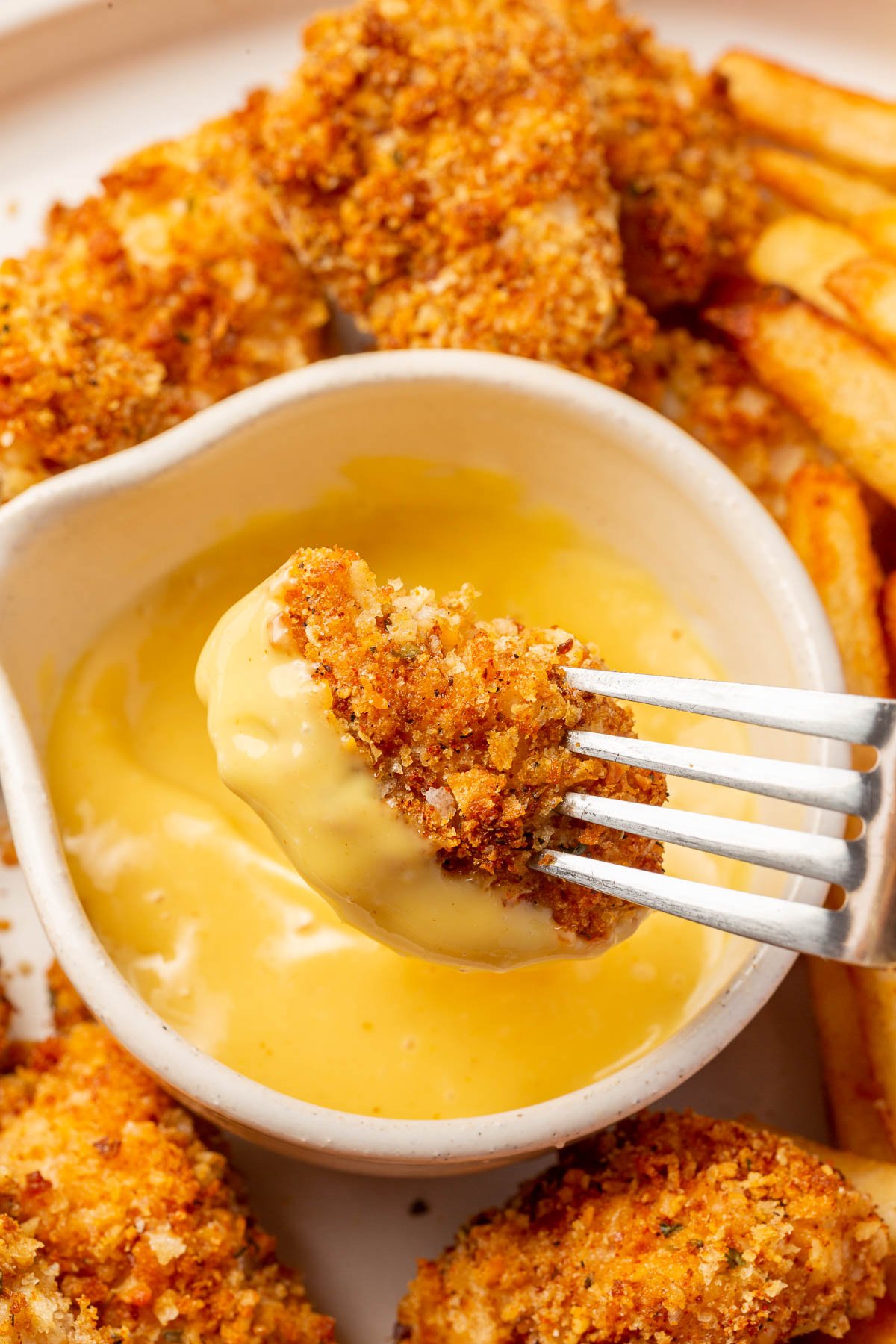 A fork dipping a gluten-free chicken nugget into a small bowl of honey mustard dipping sauce with additional gf chicken nuggets and french fries surrounding the bowl.