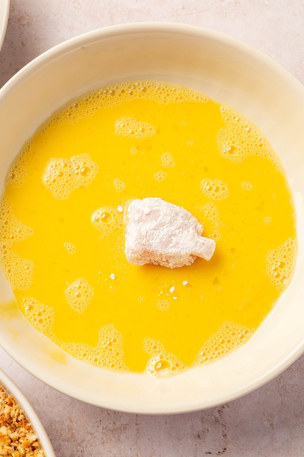 A gluten-free flour coated chicken cube in a bowl of beaten eggs.