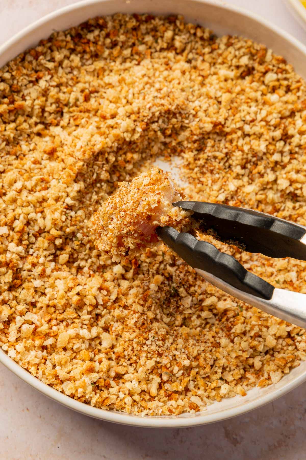 A chicken cube that has been breaded in gluten-free breadcrumbs being removed from the shallow bowl of breadcrumbs with a pair of tongs.