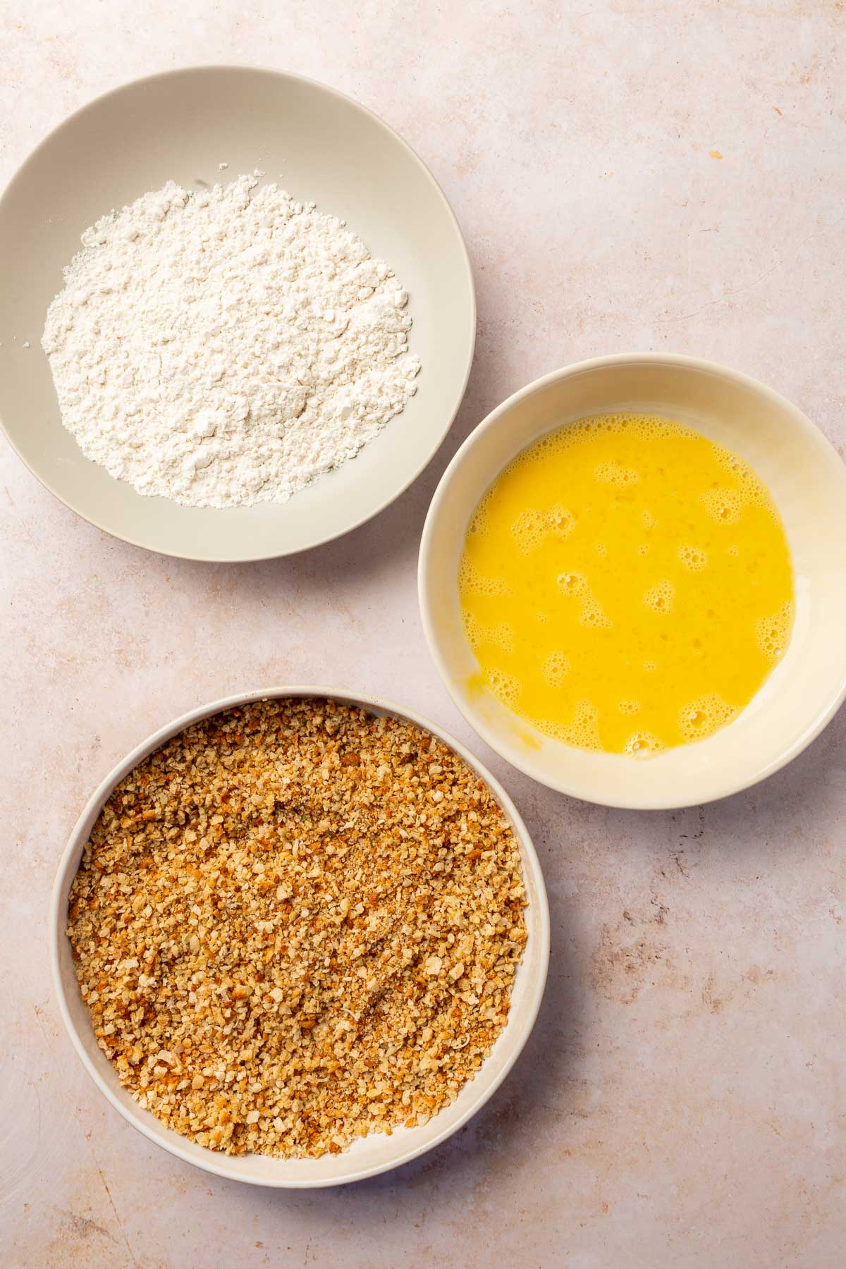 An overhead view of three shallow bowls that set up a breading station for chicken nuggets, one with gluten-free flour, one with beaten eggs and one with a mixture of breadcrumbs, spices, and parmesan cheese.