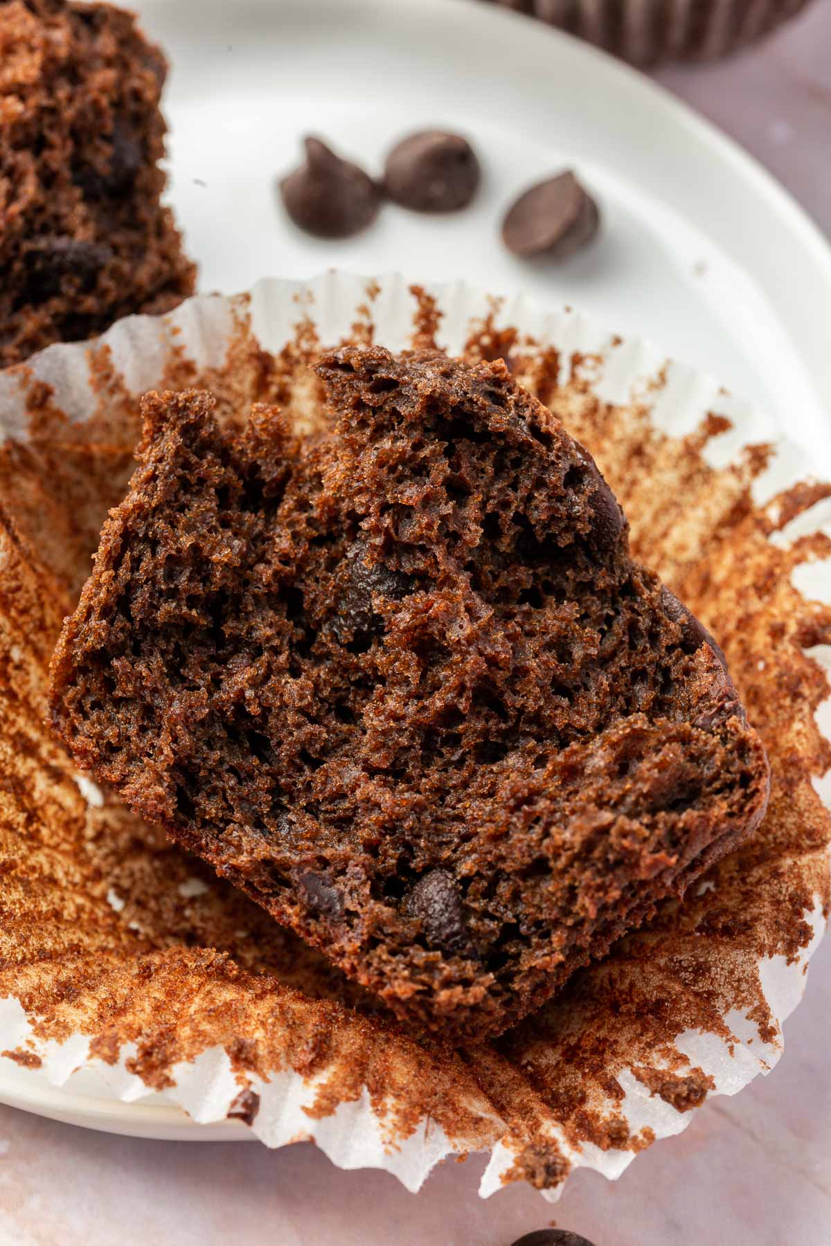 Close-up of a gluten-free chocolate muffin split in half on an unwrapped liner, showing the rich, moist texture with chocolate chips visible inside.