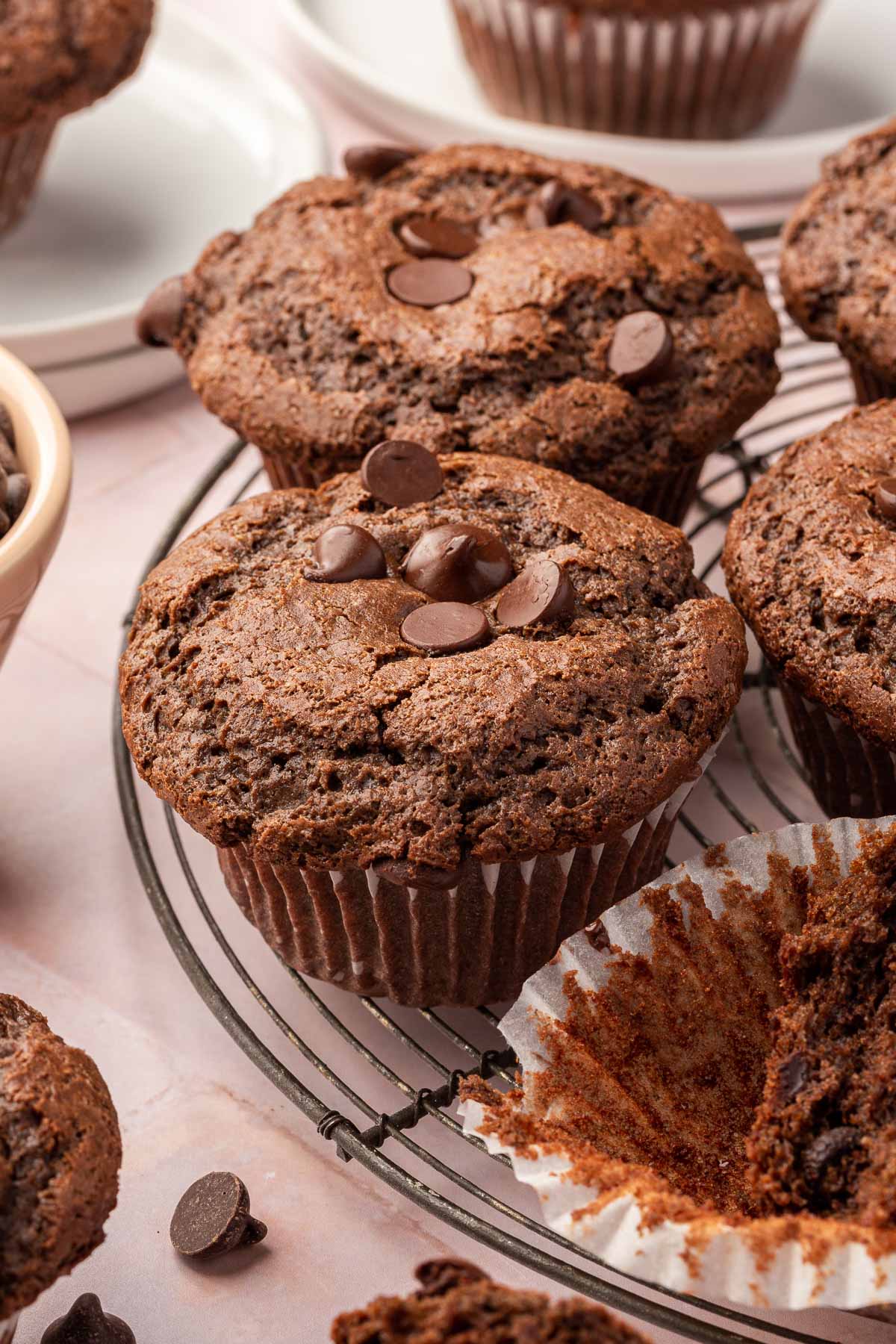 Close-up of multiple gluten-free chocolate muffins on a cooling rack, topped with chocolate chips, with one muffin partially unwrapped to show the moist interior.