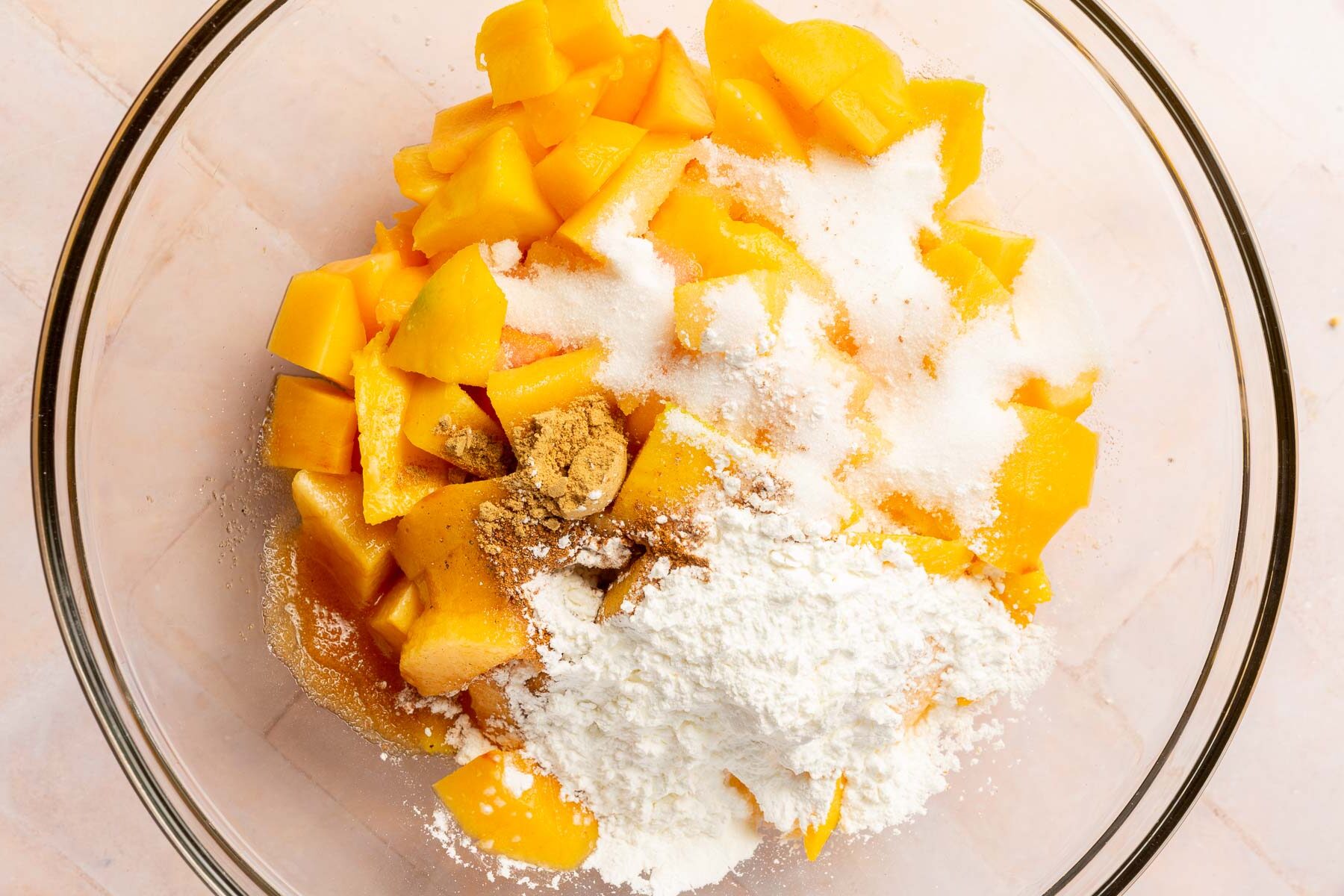 A glass mixing bowl filled with diced peaches, diced mango, granulated sugar, cornstarch, cinnamon, vanilla, water, nutmeg, and ginger before mixing together.