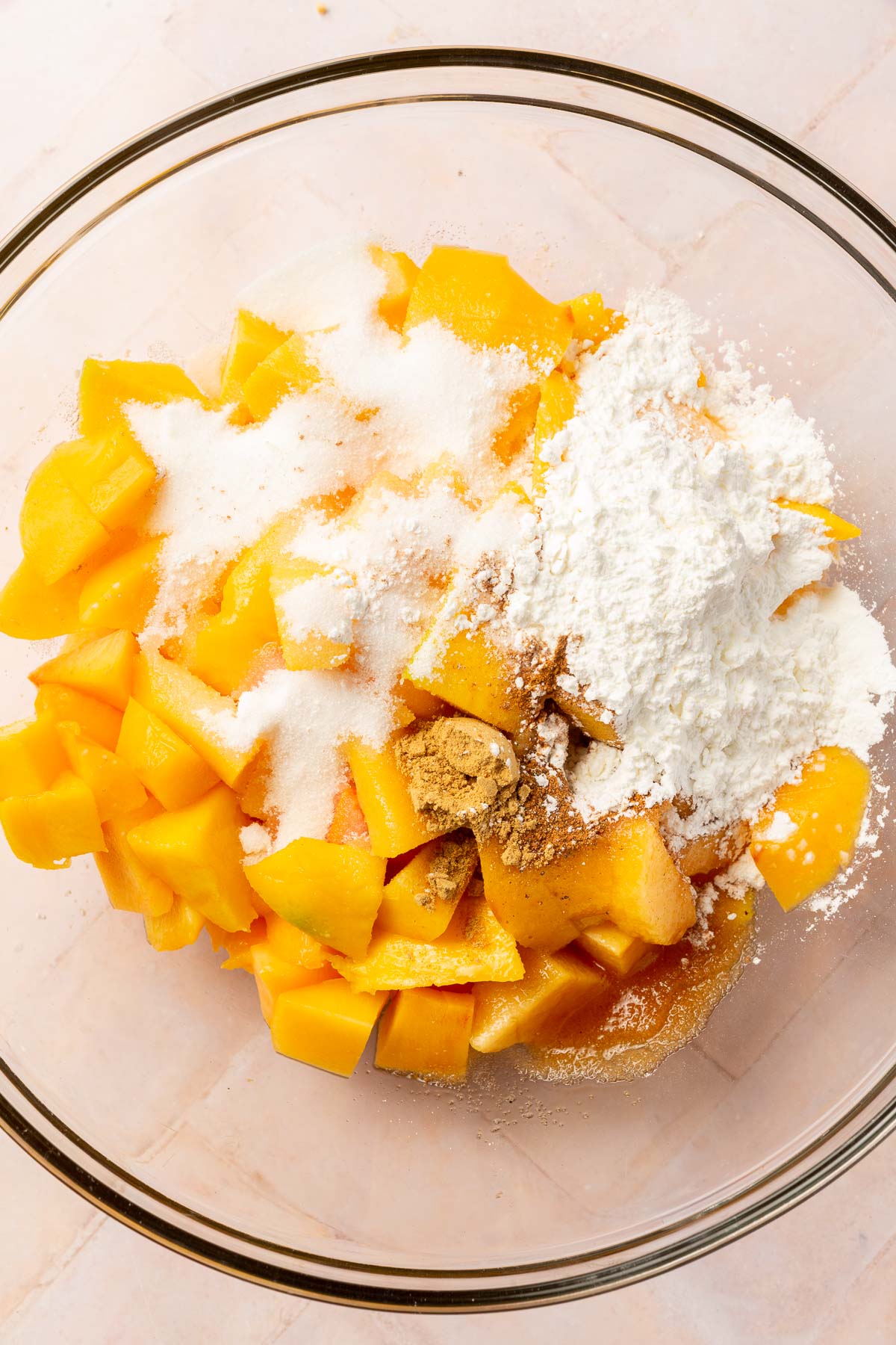 A glass mixing bowl filled with diced peaches, diced mango, granulated sugar, cornstarch, cinnamon, vanilla, water, nutmeg, and ginger before mixing together.
