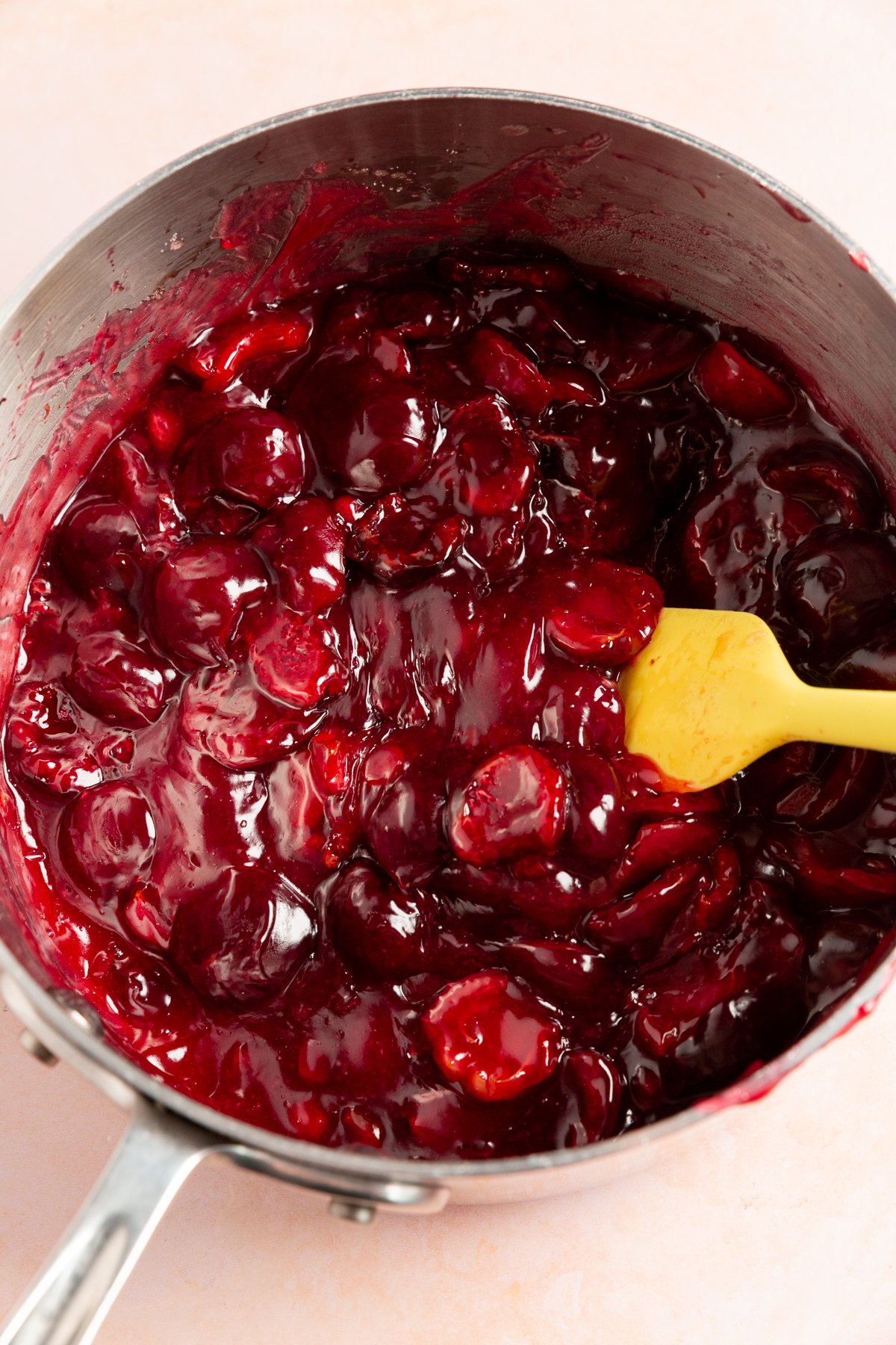 The cherry pie filling thickened and glossy, stirred with a yellow spatula in a saucepan.