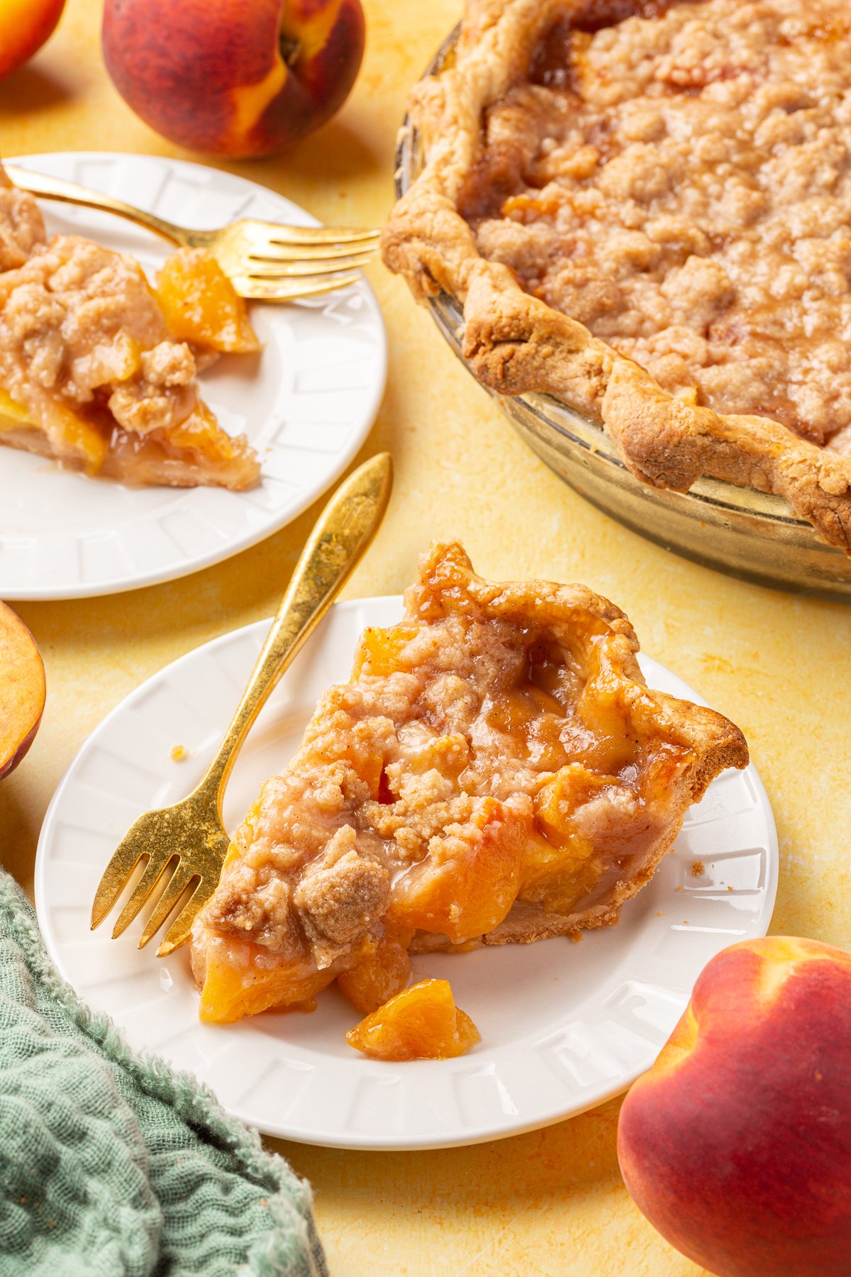 Two slices of gluten-free peach crumble pie served on white plates with gold forks, next to the pie dish and fresh peaches