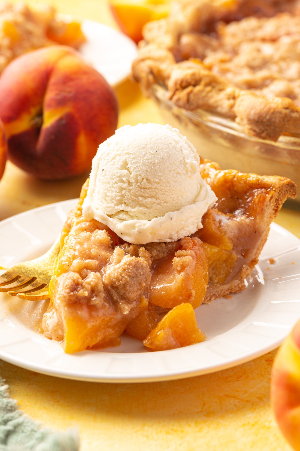 A slice of gluten-free peach crumble pie served with a scoop of vanilla ice cream on top, surrounded by whole peaches