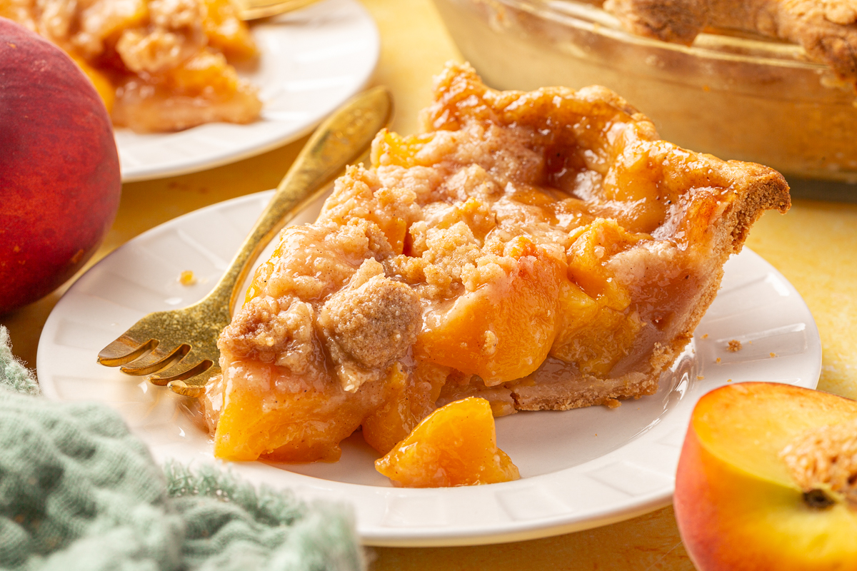 A close-up of a slice of gluten-free peach crumble pie on a white plate with a gold fork, next to a whole peach.