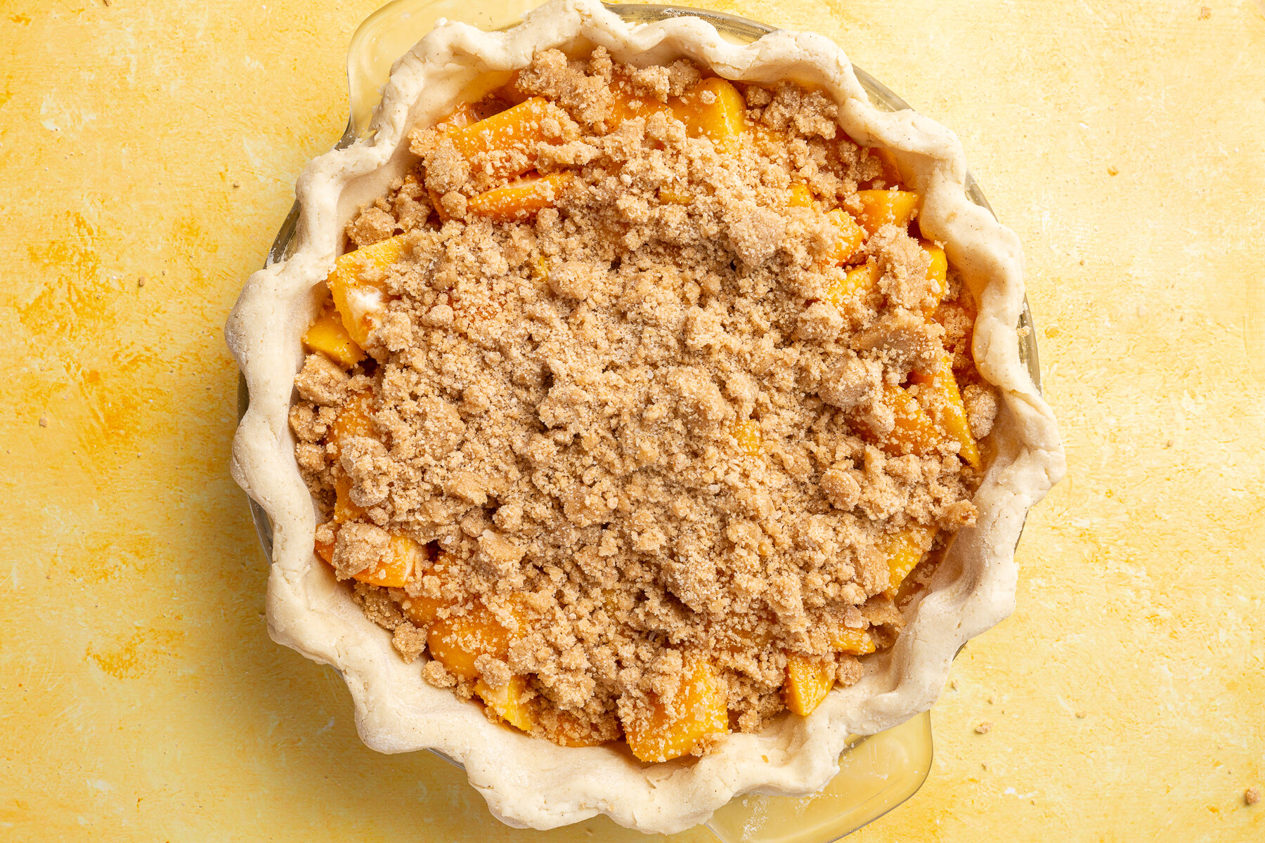 Gluten-free crumble topping spread evenly over the peach filling in the pie dish, ready to be baked.