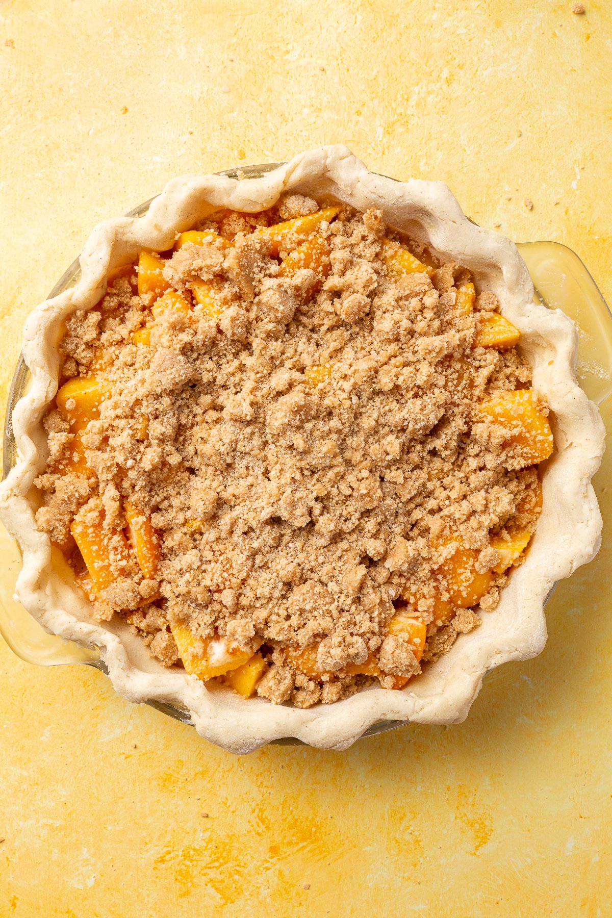 Gluten-free crumble topping spread evenly over the peach filling in the pie dish, ready to be baked.