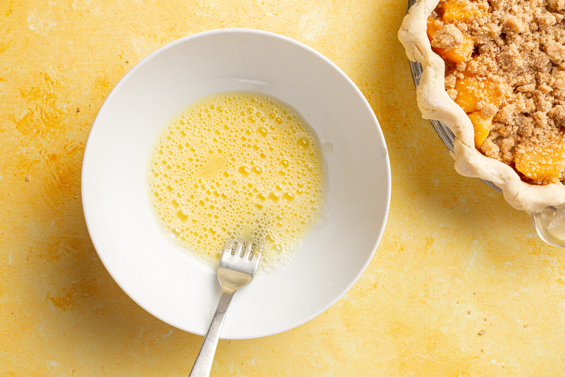 Beaten egg in a white bowl next to an unbaked peach crumble pie.