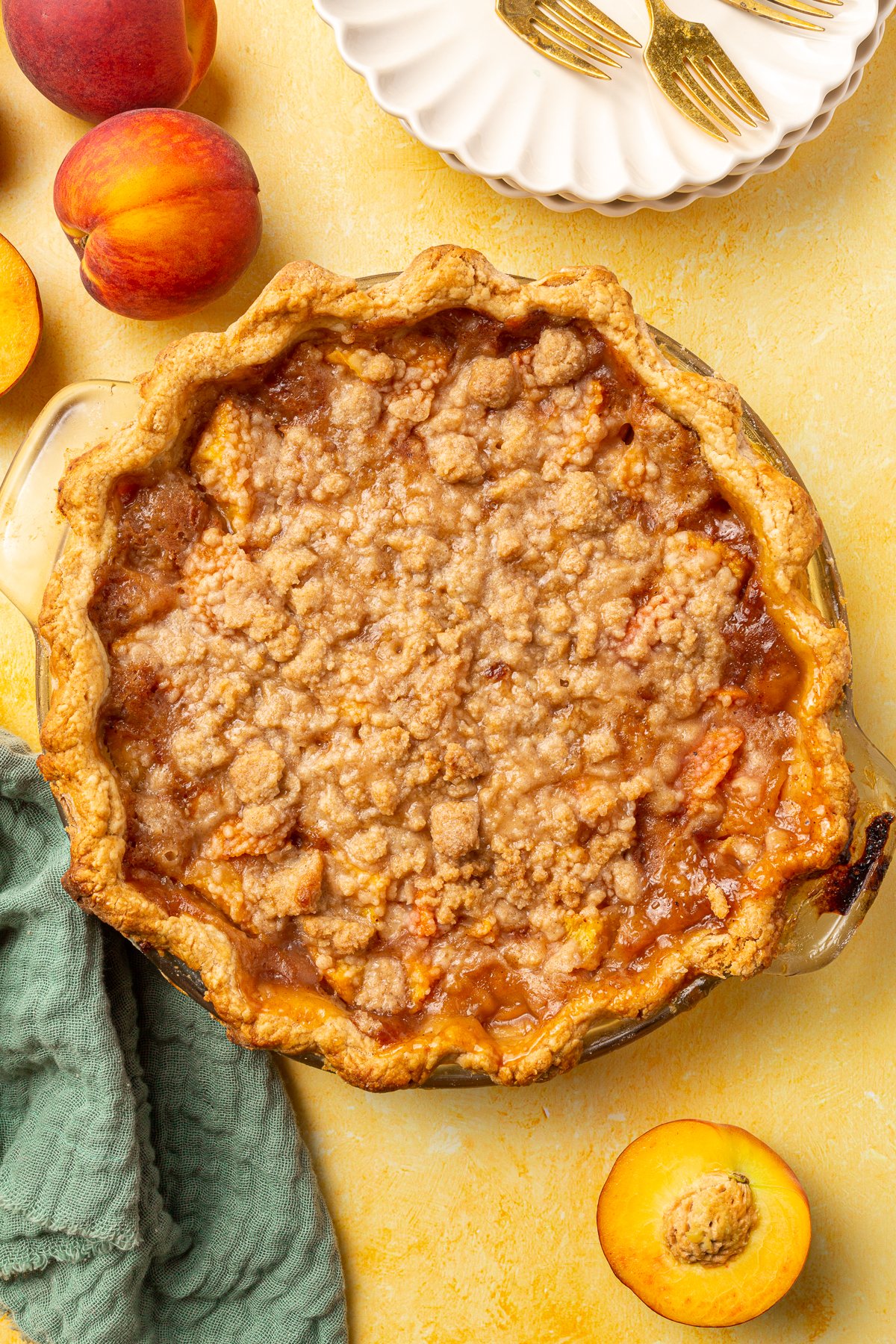 Baked peach crumble pie next to fresh peaches, a plate with forks, and a green napkin.