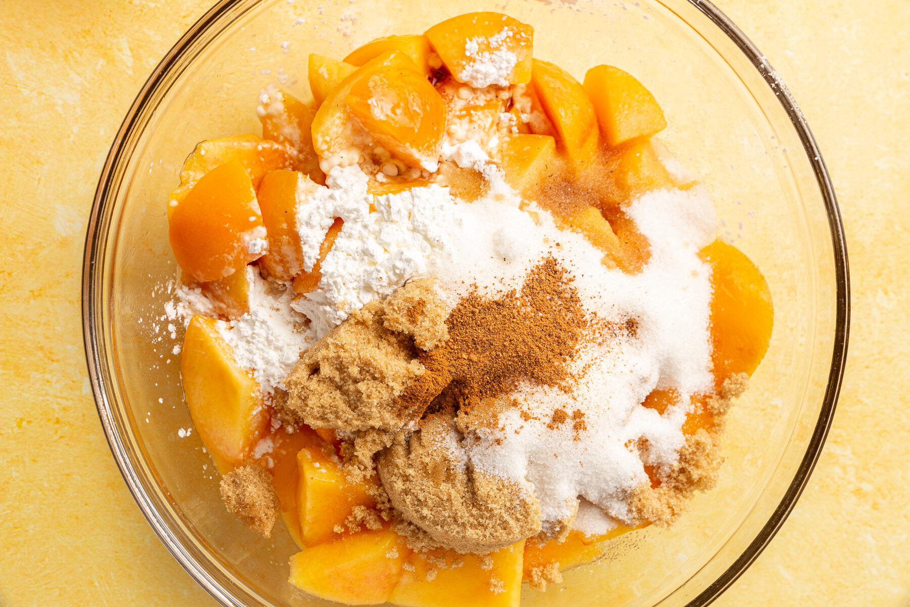Chopped peaches topped with sugar, brown sugar, flour, and cinnamon in a glass bowl, ready to be mixed.