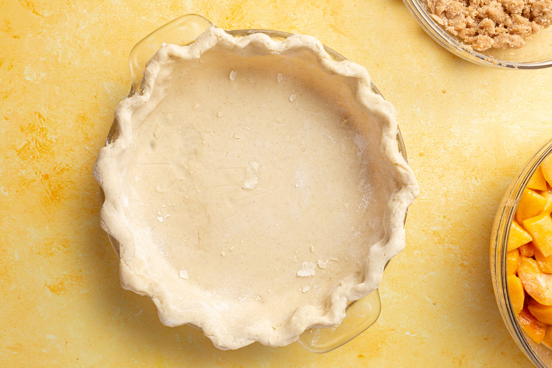Unbaked gluten-free pie crust pressed into a pie dish, with bowls of peach filling and crumble topping in the background.