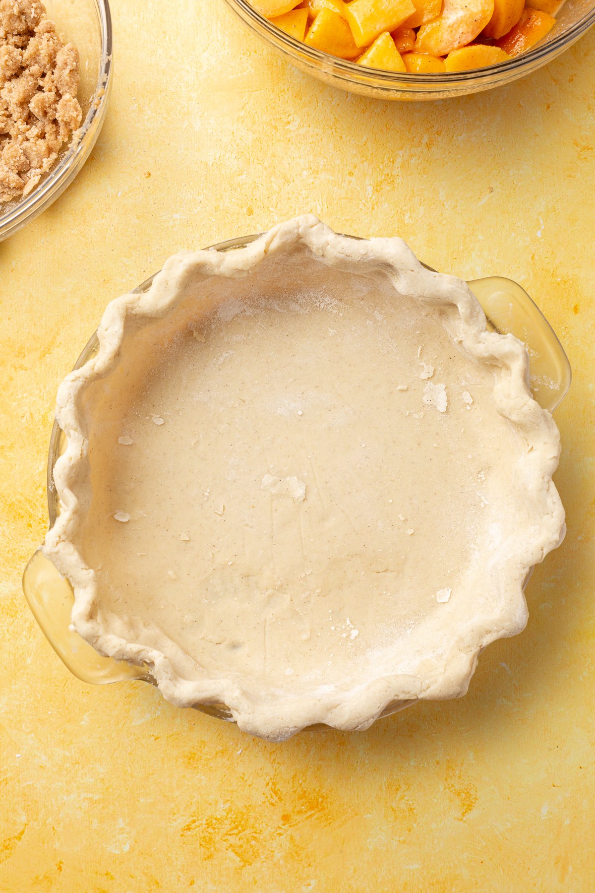 Unbaked gluten-free pie crust pressed into a pie dish, with bowls of peach filling and crumble topping in the background.