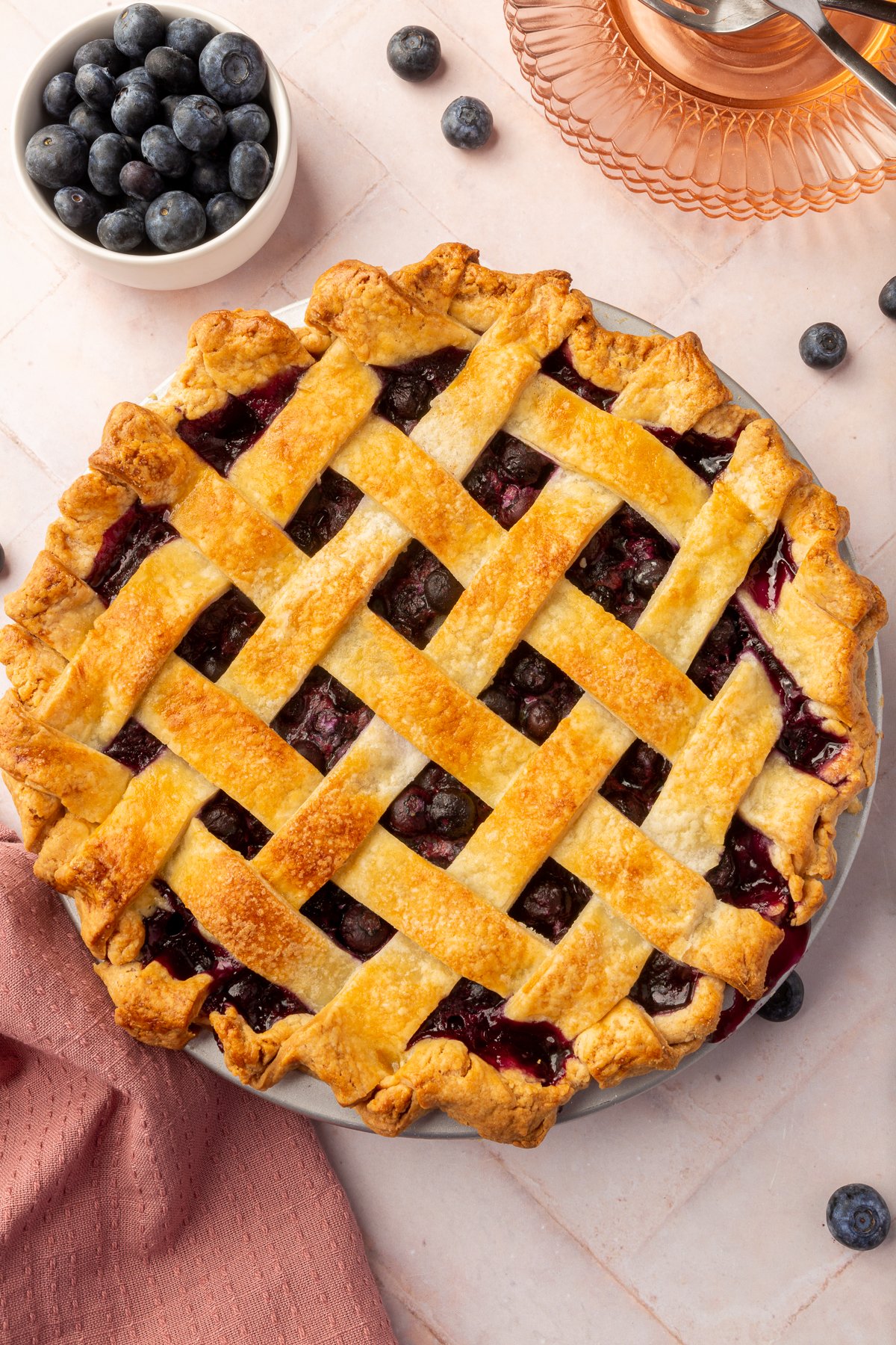 An overhead shot of a whole blueberry pie with a lattice crust. A small bowl of blueberries is placed next to the pie.