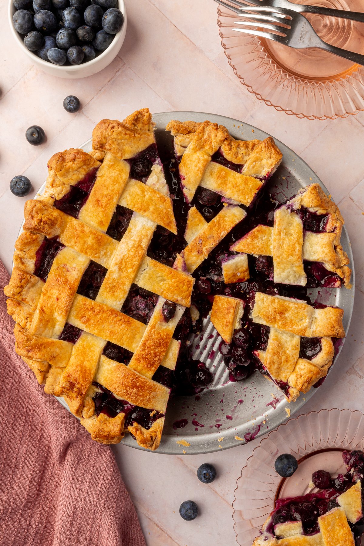 A partially sliced blueberry pie with one slice on a glass plate nearby and fresh blueberries scattered around the scene.