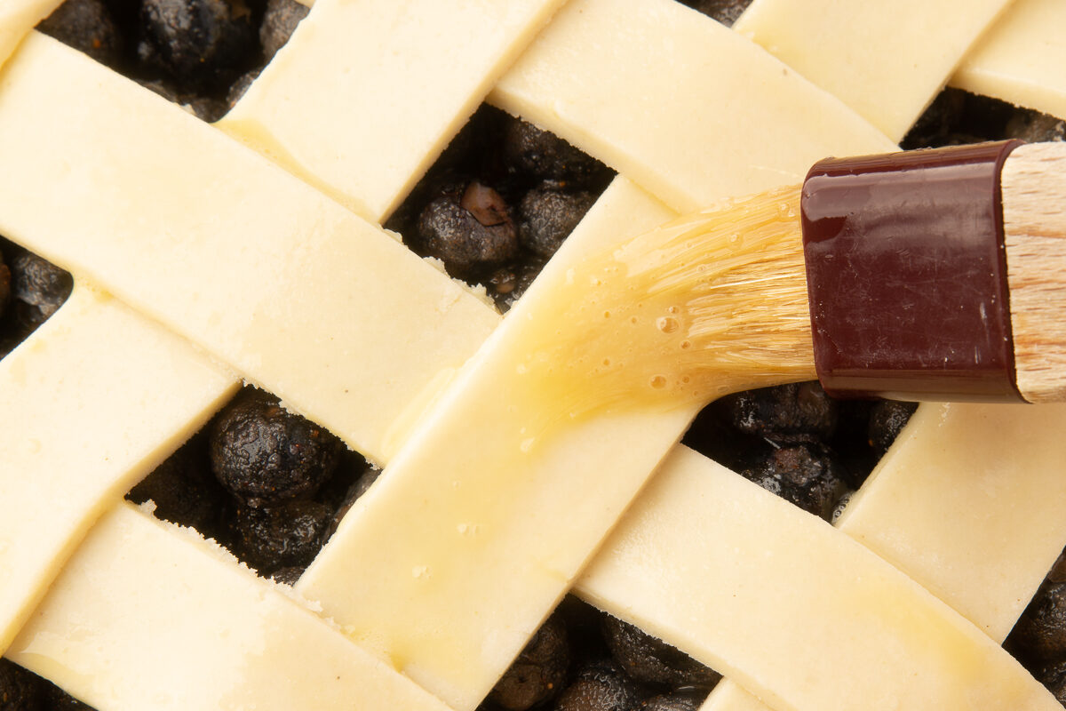 A pastry brush applying egg wash to the lattice top of the blueberry pie to create a golden crust.