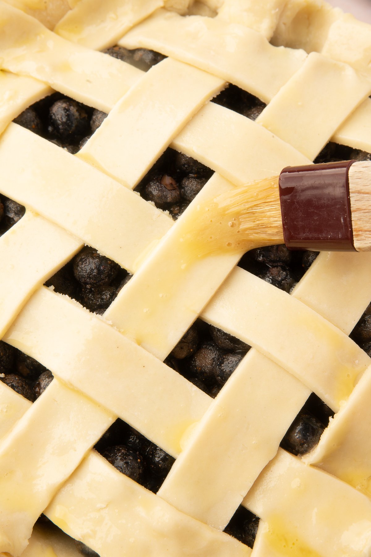 A pastry brush applying egg wash to the lattice top of the blueberry pie to create a golden crust.