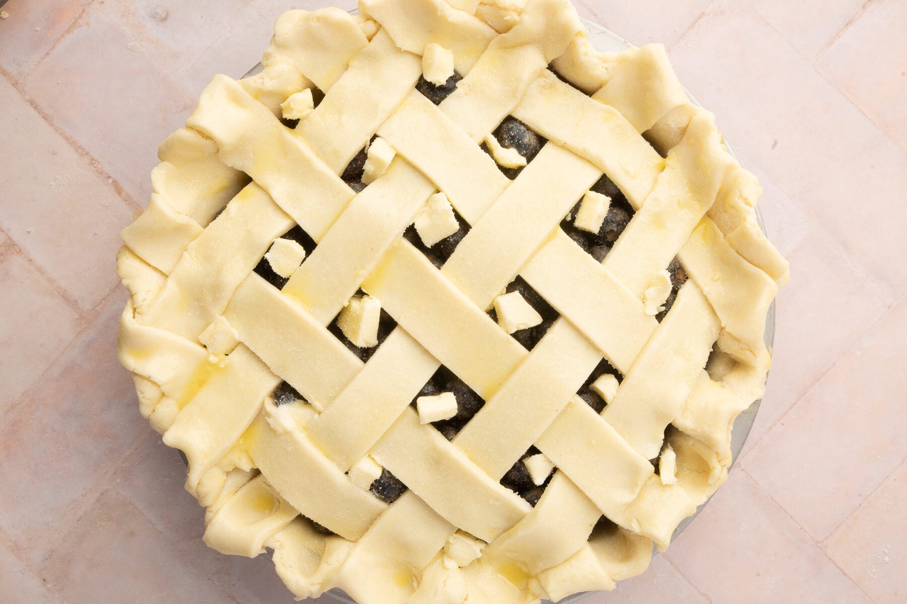 Overhead view of a blueberry pie with a lattice crust, dotted with small cubes of butter, ready to be baked. The pie crust edges are crimped, and the filling is visible through the lattice.