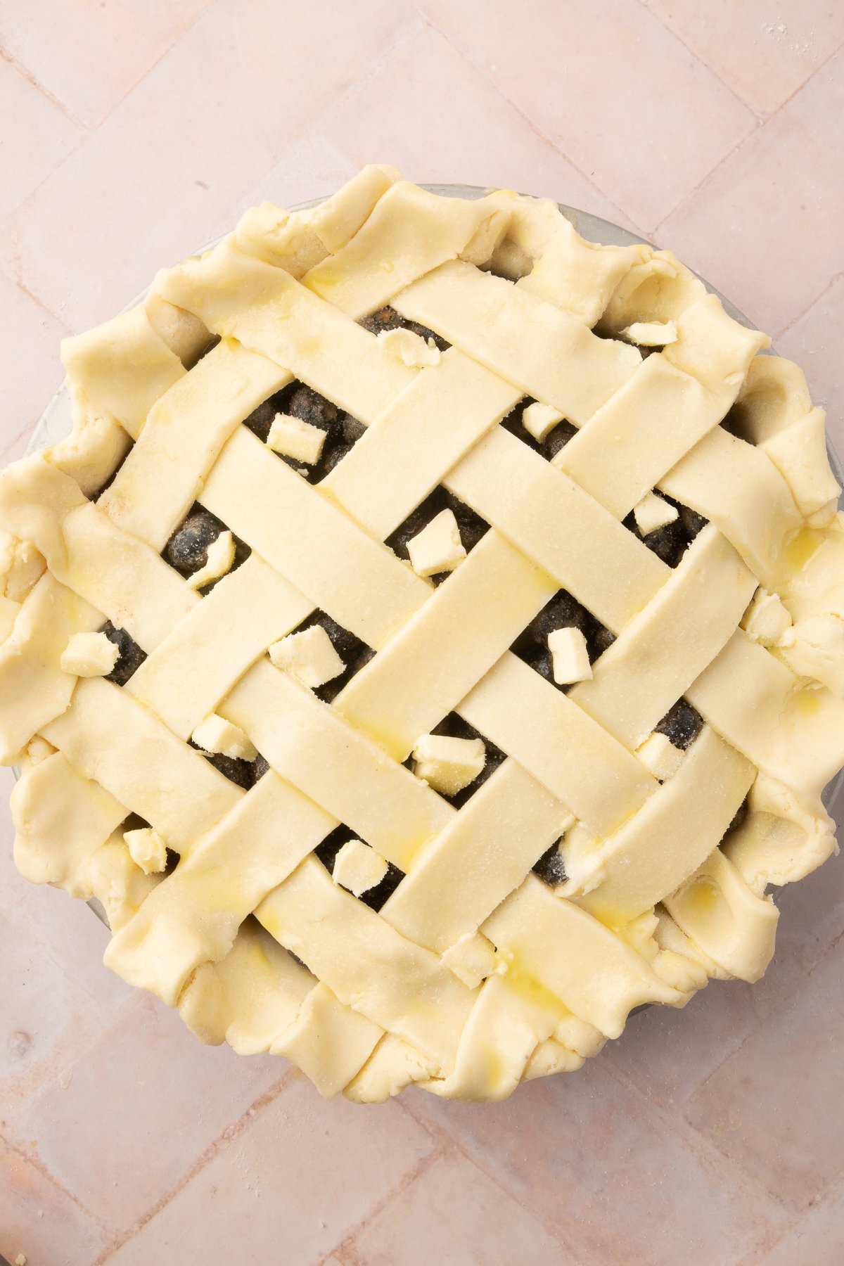 Overhead view of a blueberry pie with a lattice crust, dotted with small cubes of butter, ready to be baked. The pie crust edges are crimped, and the filling is visible through the lattice.