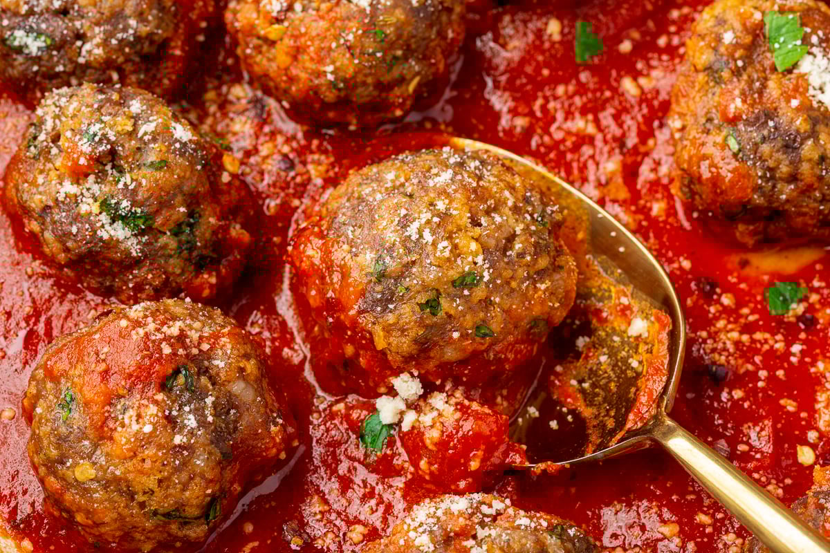Close up view of spicy meatballs made with ground beef, Italian sausage and gluten-free breadcrumbs resting in a red marinara sauce and being scooped out with a gold spoon.
