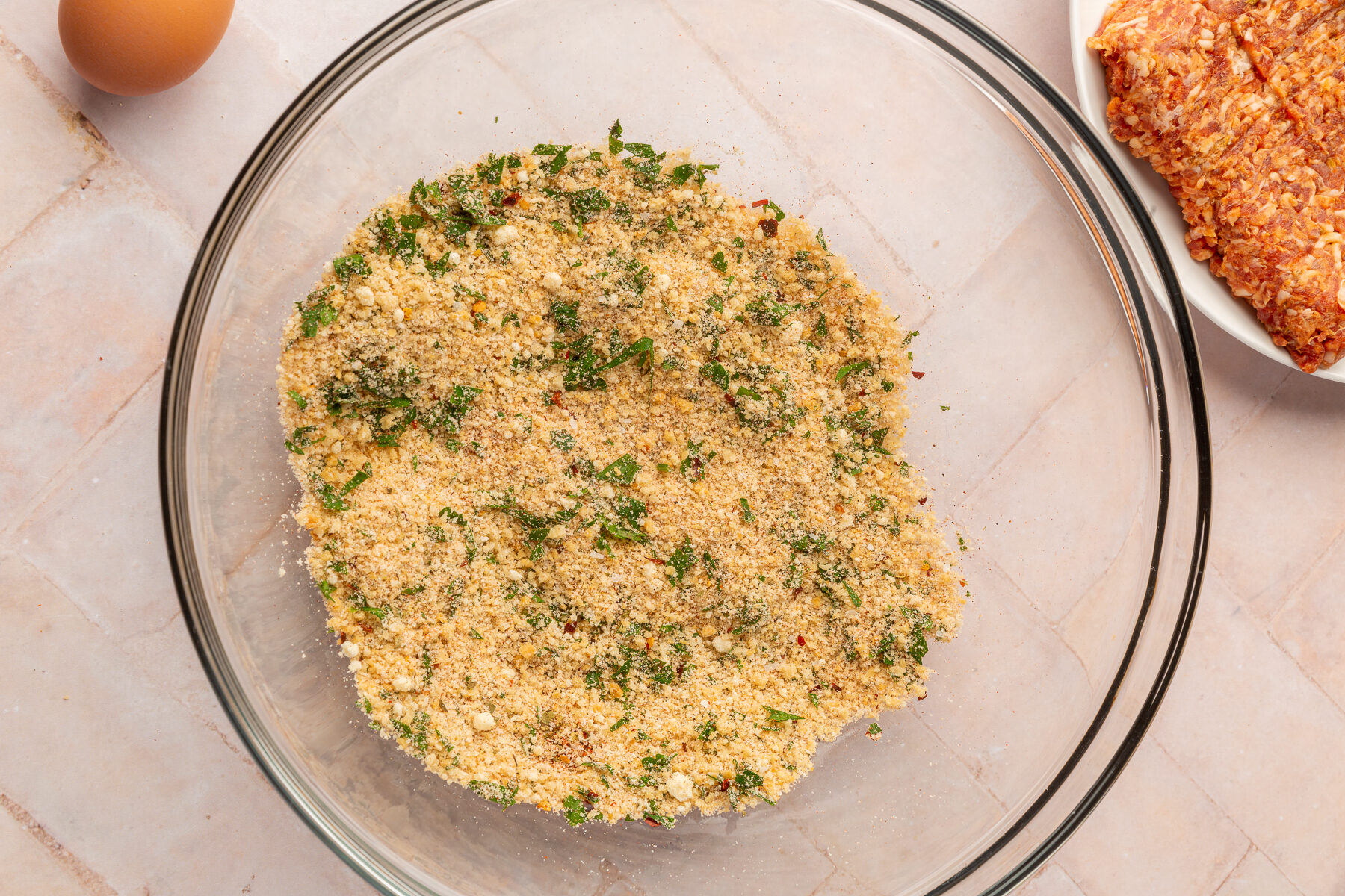 Gluten-free breadcrumbs, parmesan cheese, fresh parsley, dried oregano, garlic powder, onion powder, ground black pepper, salt, red pepper flakes and paprika mixed together in a large mixing bowl on a brick countertop.