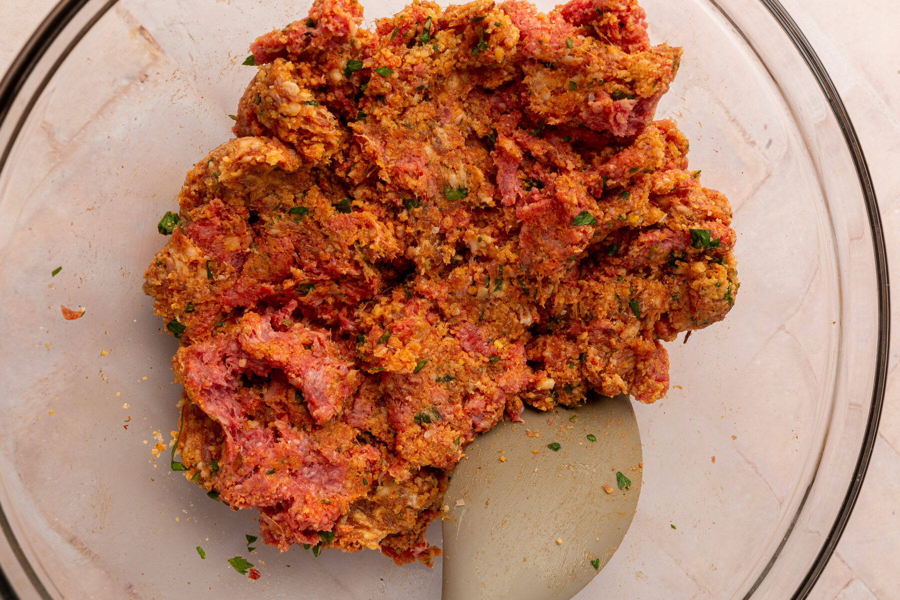 Ingredients for spicy meatballs mixed together in a large mixing bowl, including ground beef, Italian sausage, egg, gluten-free breadcrumbs, parmesan cheese, fresh parsley, dried oregano, garlic powder, onion powder, paprika, salt, ground black pepper and red pepper flakes.