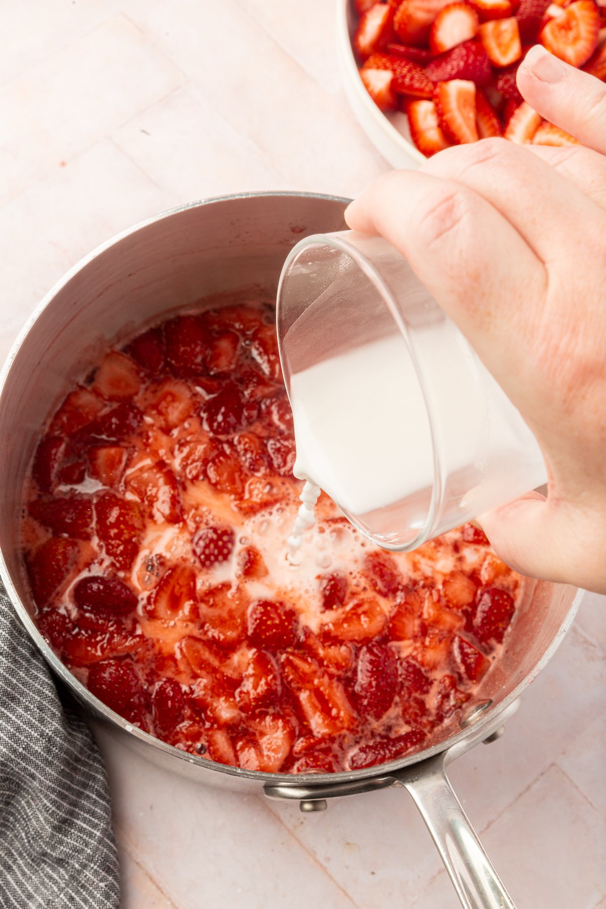 A hand pouring a white cornstarch slurry into a pot of simmering strawberries to thicken the mixture.