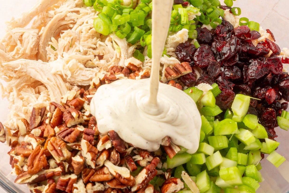 Mayonnaise, lemon juice, dijon mustard, salt and pepper mixture being poured over cooked chicken, green onions, celery, dried cranberries and pecans in a large mixing bowl.