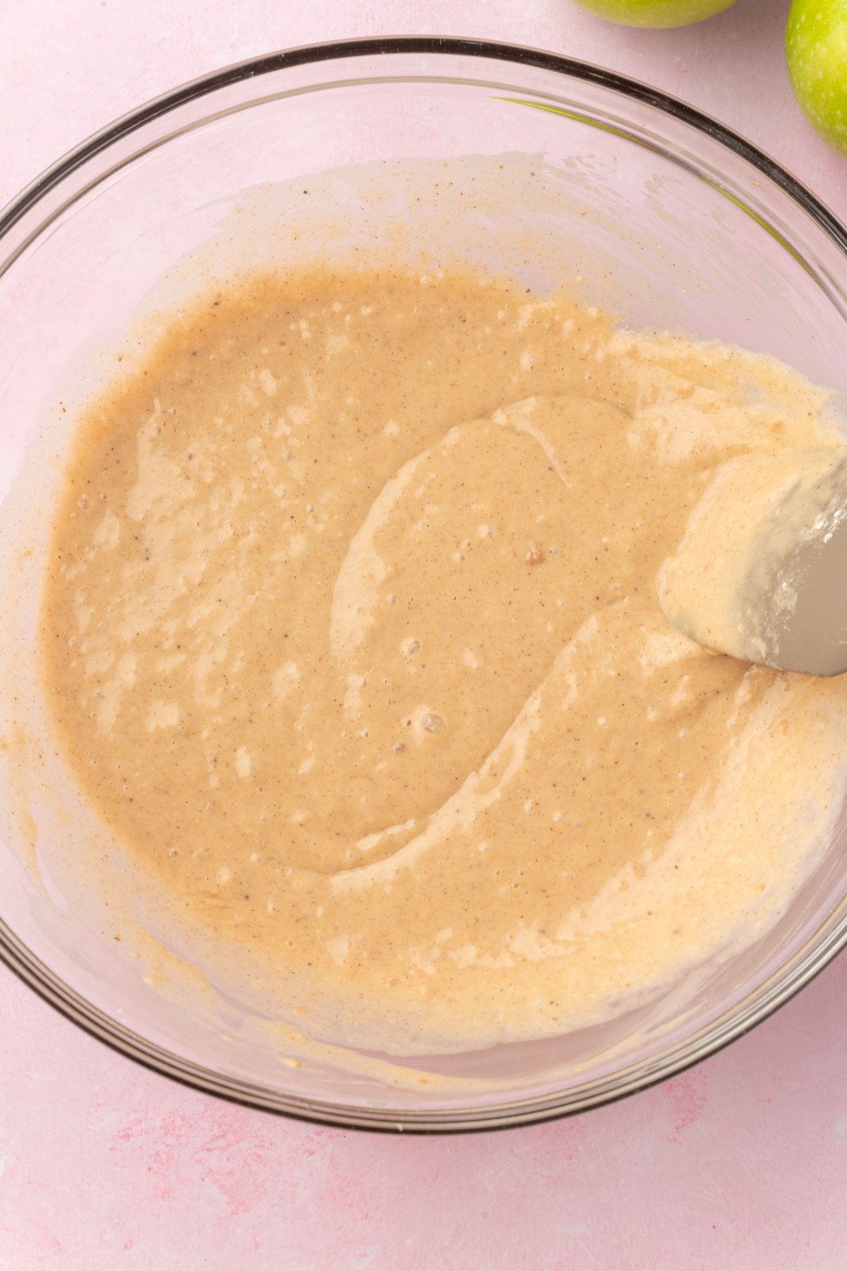 A glass mixing bowl of apple fritter batter being mixed with a silicone spatula.