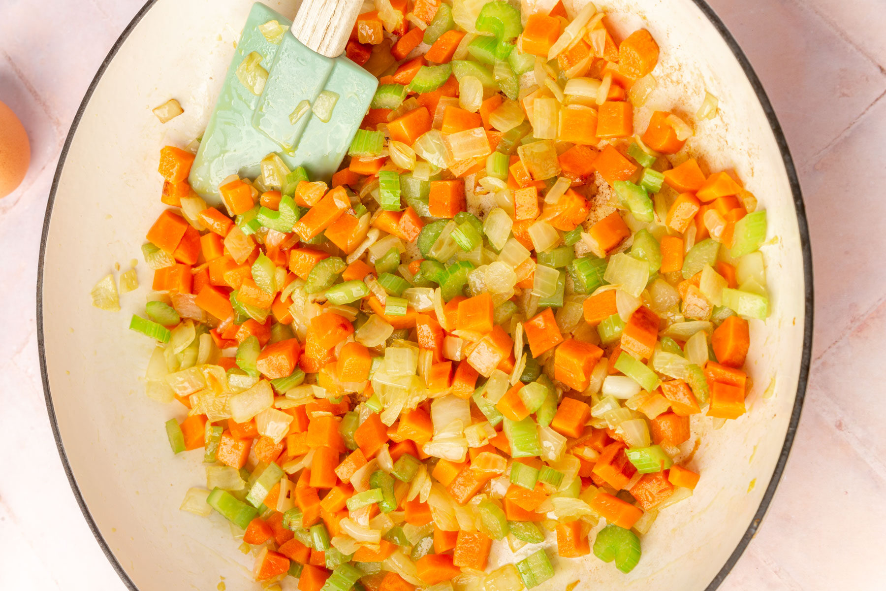 Chopped carrots, celery, and onions sautéing in a white pan with a green spatula on the side.