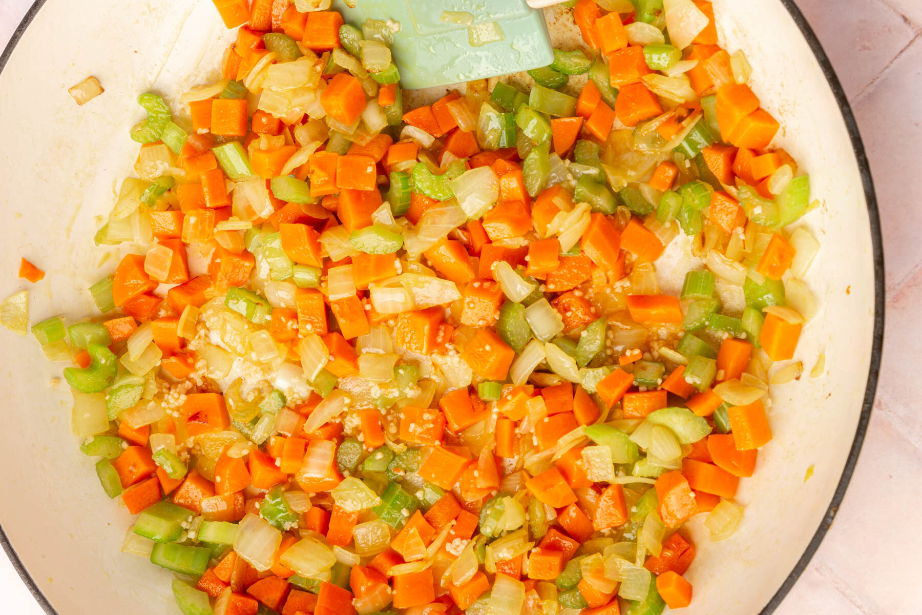 Diced carrots, celery, and onions being sautéed in a pan with a green spatula, creating a colorful vegetable mixture.
