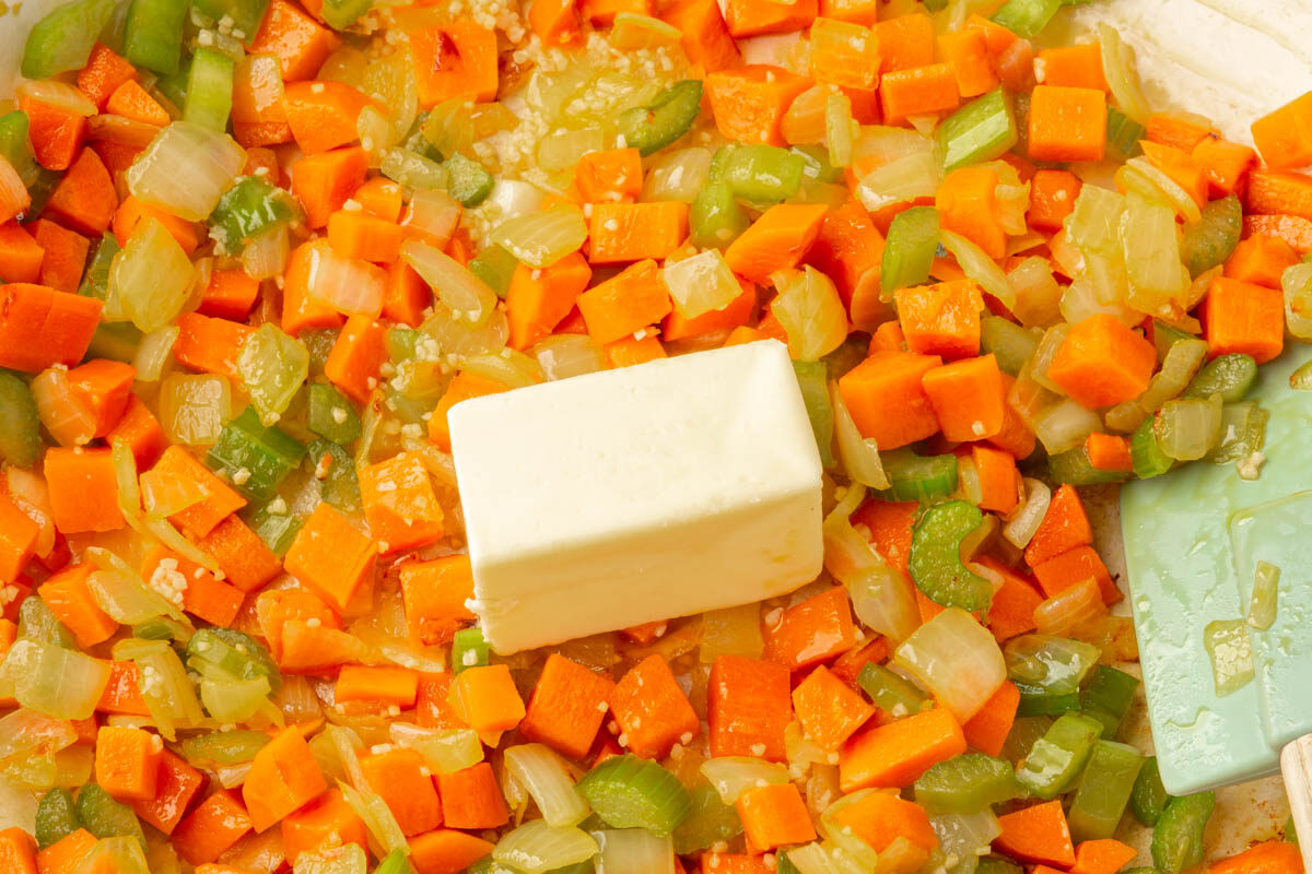 Chopped carrots, celery, and onions are sautéing in a pan with a stick of butter in the center, next to a light green spatula.