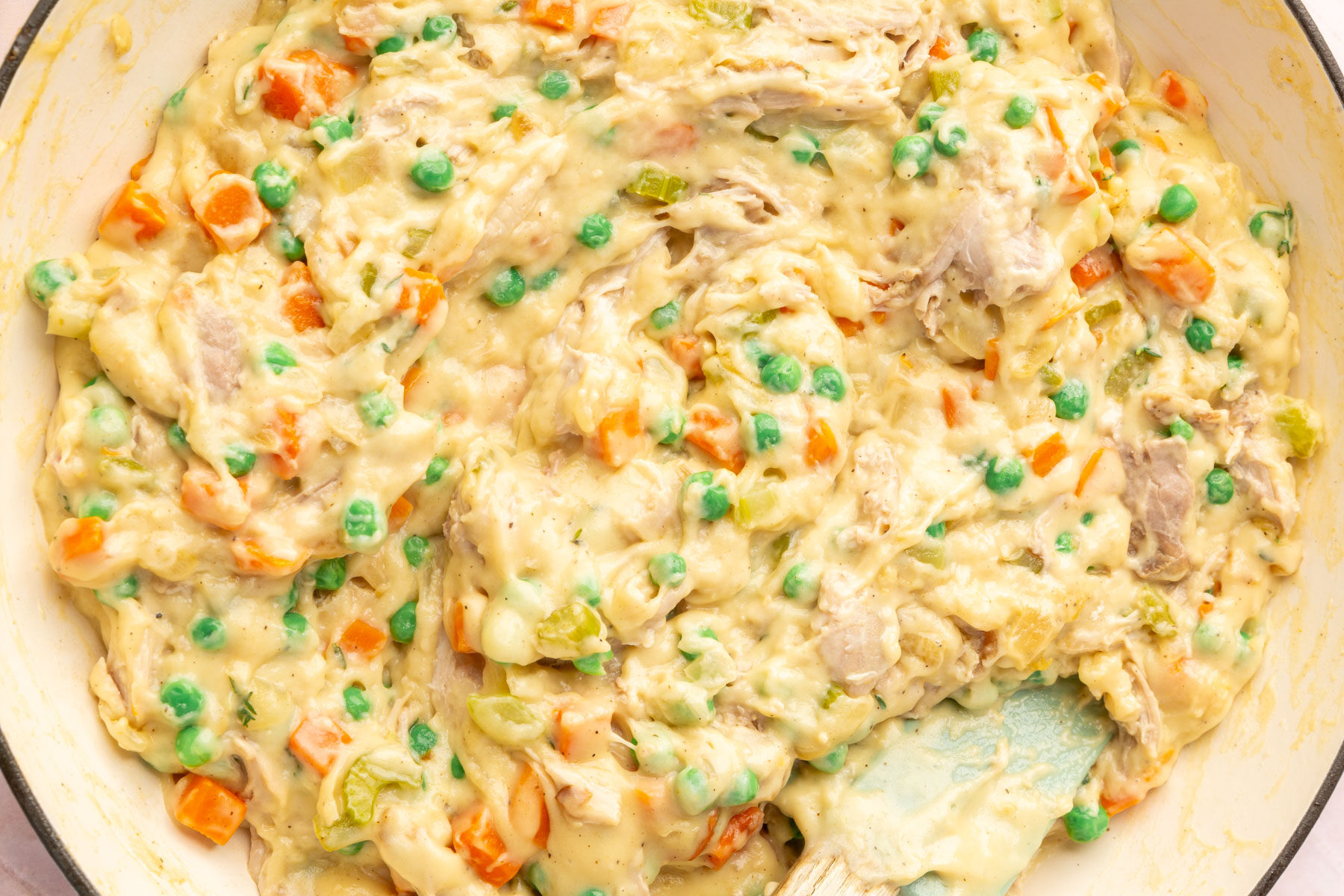 A close-up of creamy chicken pot pie filling with chunks of chicken, carrots, peas, celery, and a thick sauce, being mixed in a white bowl with a spatula.