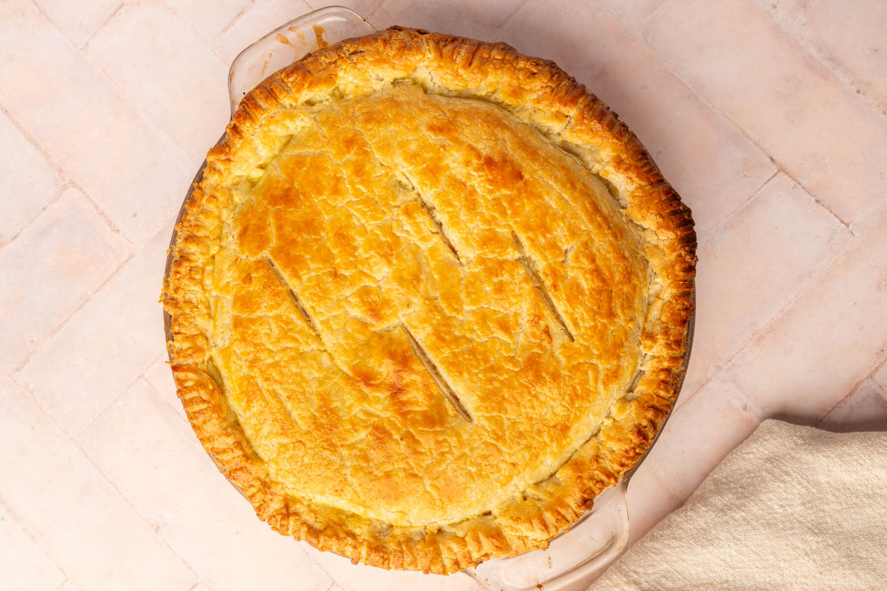A golden-brown pie with a flaky crust sits in a glass pie dish on a light-colored tiled surface, with a beige cloth partially visible in the bottom right corner.