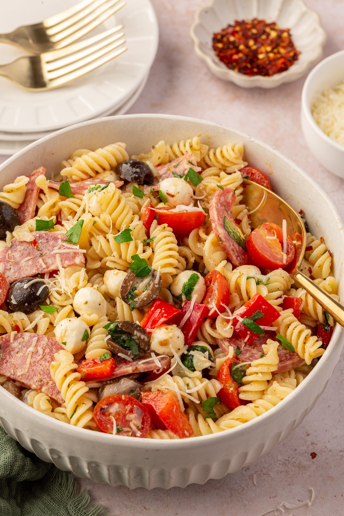 Photo of large serving bowl of gluten-free pasta salad with large serving spoon on table with various ingredients in the background.