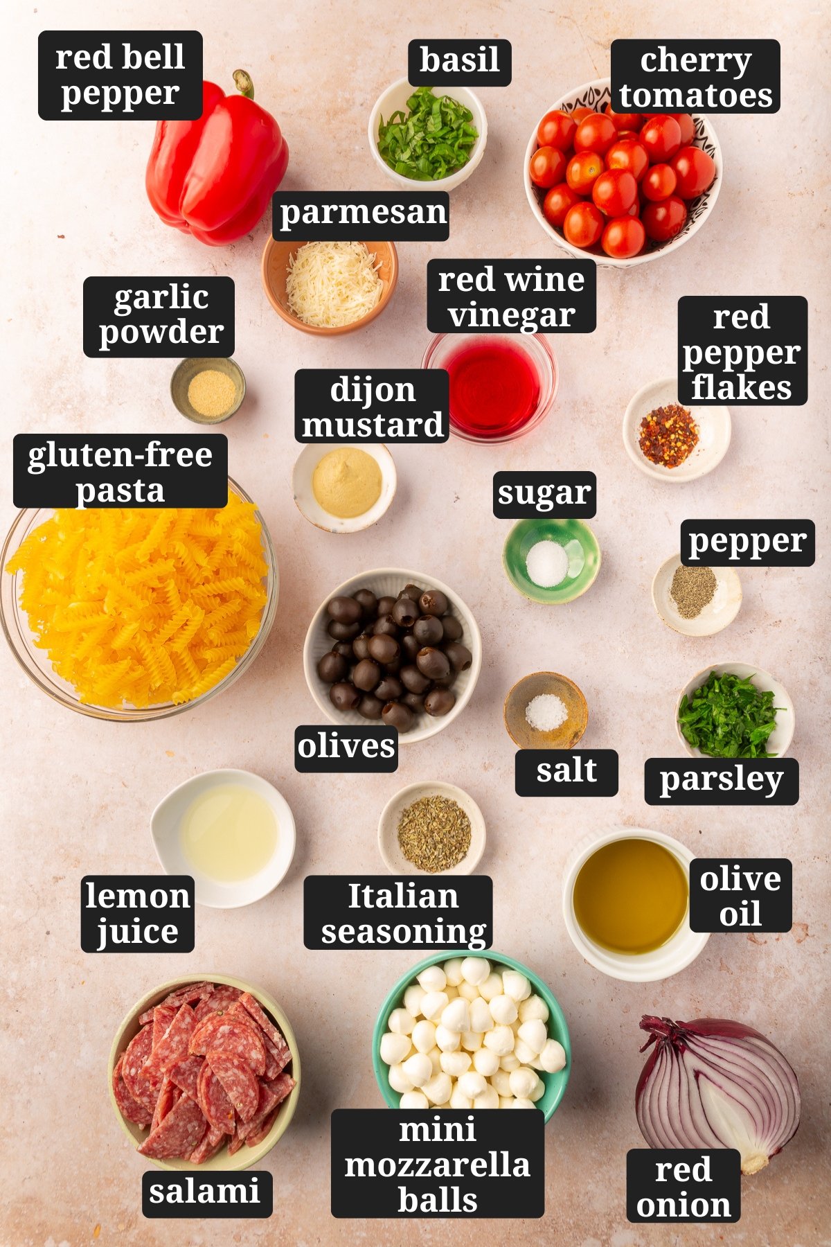 Ingredients for gluten-free pasta salad all in individual bowls and labeled including red bell pepper, basil, cherry tomatoes, garlic powder, parmesan, red wine vinegar, red pepper flakes, sugar, pepper, dijon mustard, gluten-free pasta, olives, salt, parsley, olive oil, Italian dressing, lemon juice, salami, mini mozzarella balls, and red onion.