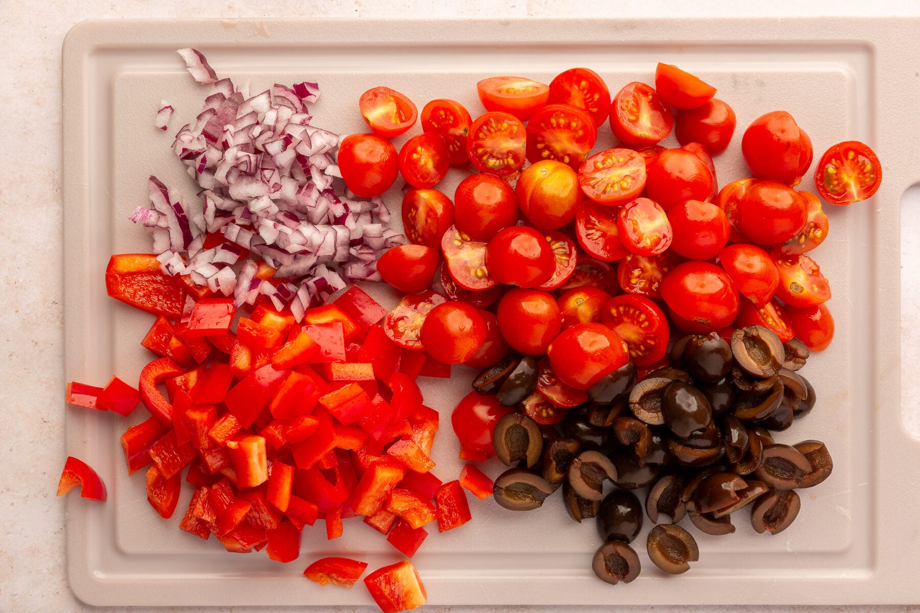 Cherry tomatoes, olives, red peppers and red onions, all chopped on a beige cutting board.