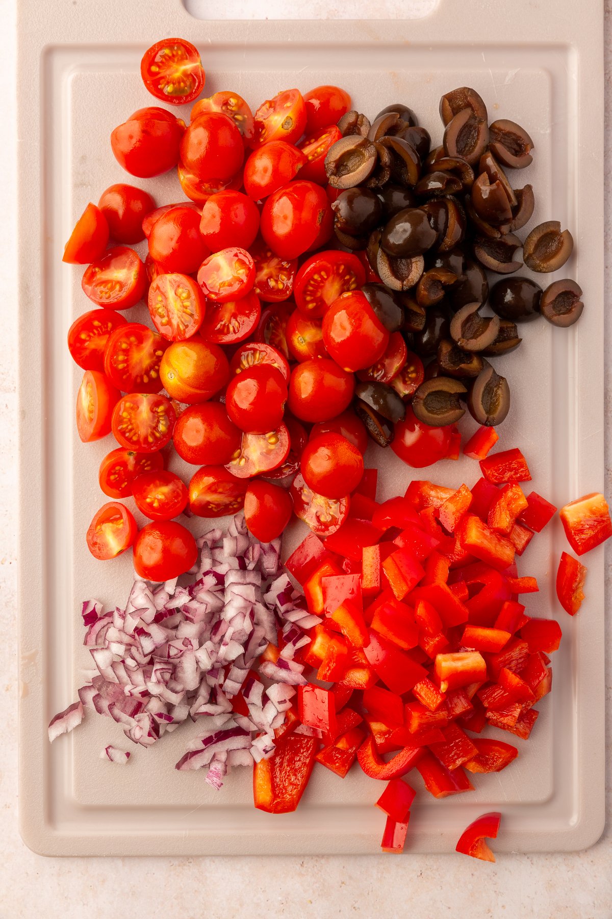 Cherry tomatoes, olives, red peppers and red onions, all chopped on a beige cutting board.
