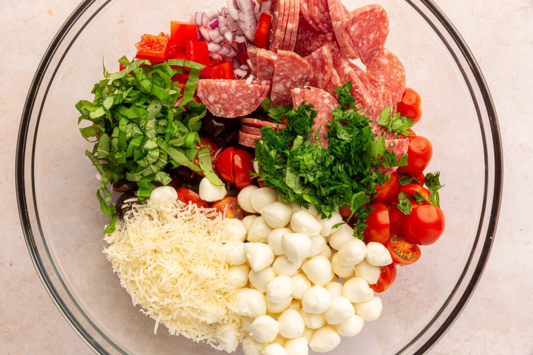 Mozzarella balls, cherry tomatoes, basil, parsley, parmesan cheese, red pepper, red onion, olives, and salami in a large mixing bowl, not yet mixed, on a marble countertop.