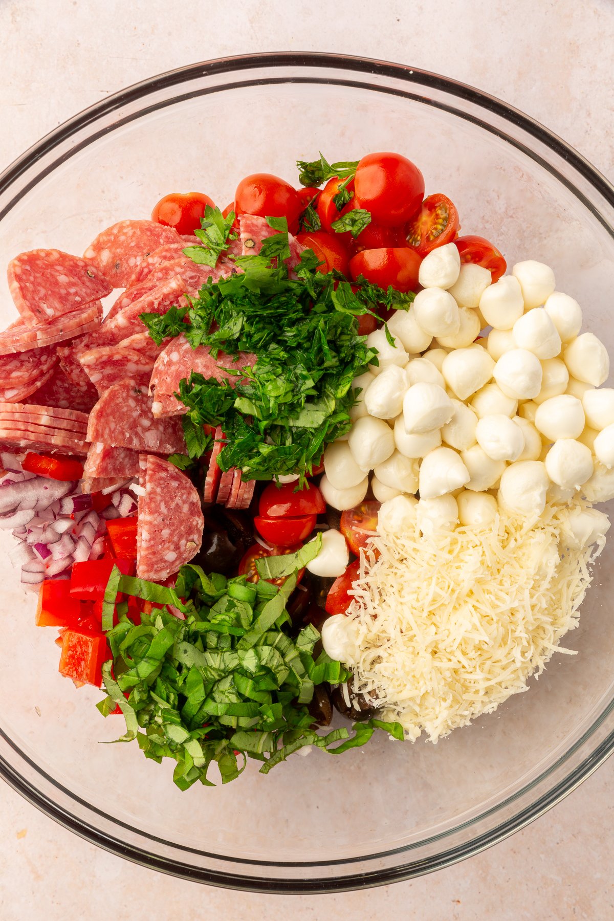 Mozzarella balls, cherry tomatoes, basil, parsley, parmesan cheese, red pepper, red onion, olives, and salami in a large mixing bowl, not yet mixed, on a marble countertop.